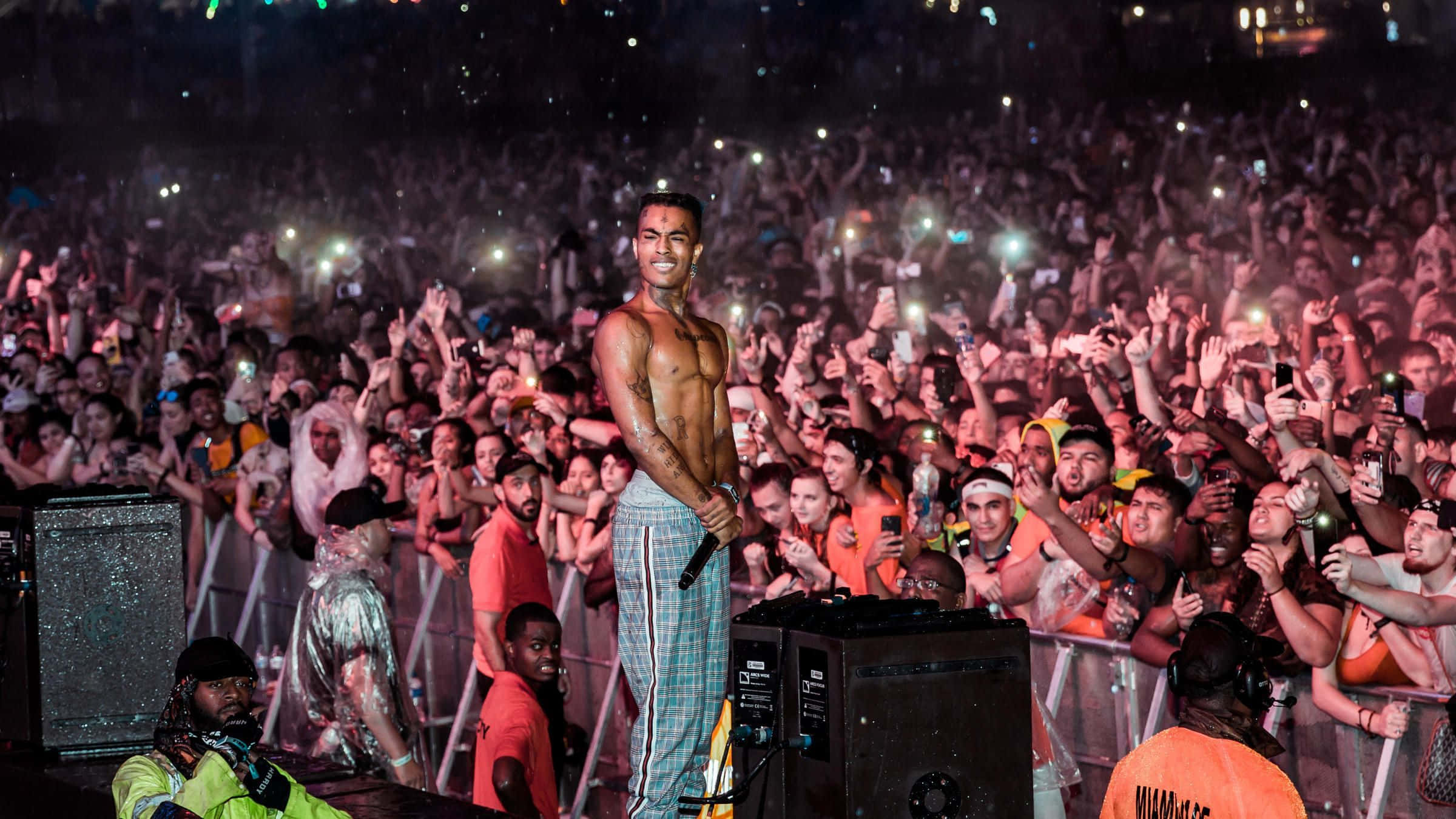 XXXTENTACION 桌布 壁紙 — 一位赤裸上身的表演者在夜間的舞台上咧嘴笑，而大量人群在濕透的音樂會中舉起發光的手機。