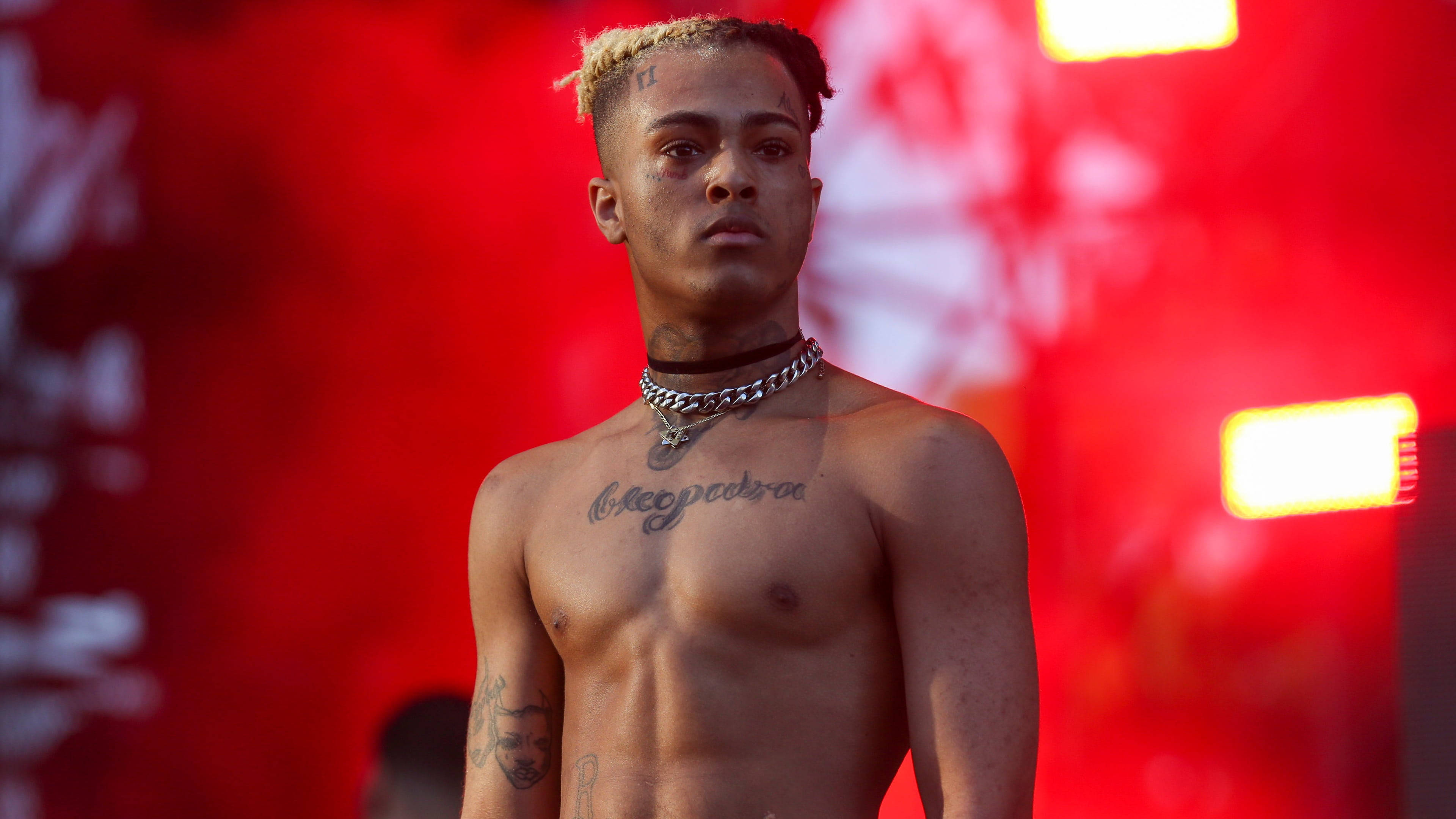xxxtentacion 電腦 桌布 壁紙 — 一位赤裸上身的金髮刺青年輕男子，自信地站在充滿活力的紅色舞台背景前，周圍有發光的燈光。