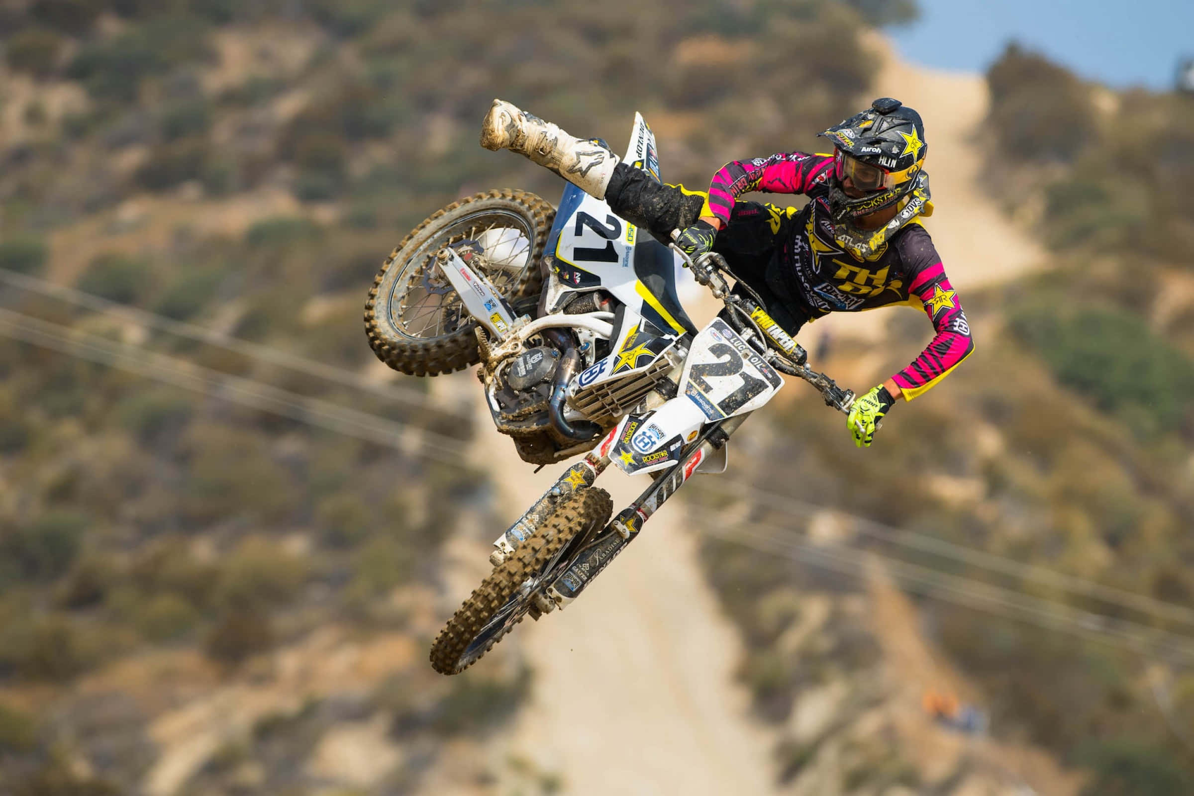 Motocross 桌布 壁紙 — 一位越野摩托車手在崎嶇的泥地摩托車上飛躍空中，背景是陽光照耀的山坡。