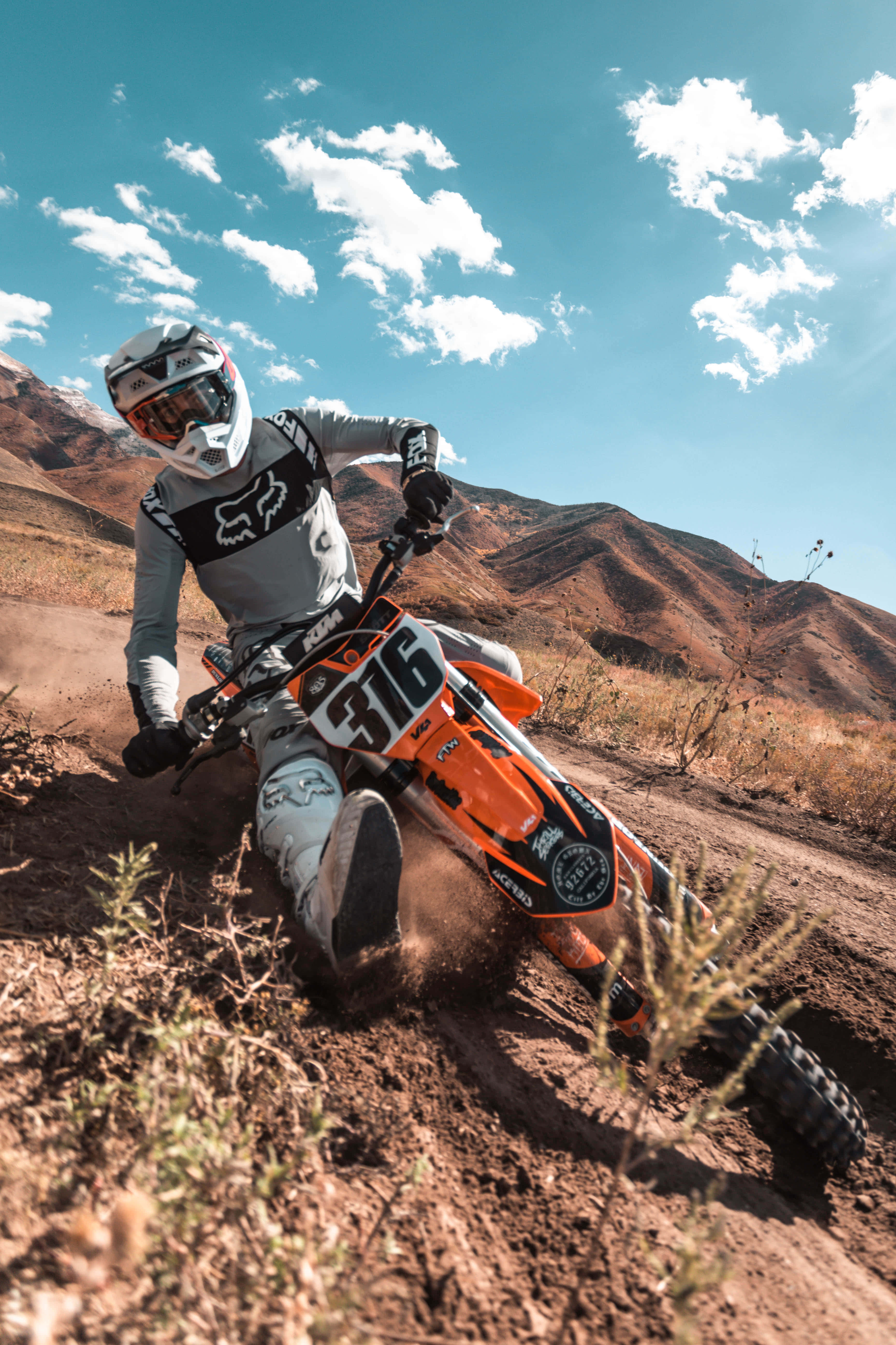 Motocross 桌布 壁紙 — 一位越野摩托車手在橙色越野車上傾斜入彎，揚起塵土，背景是乾燥的山脈和藍天。