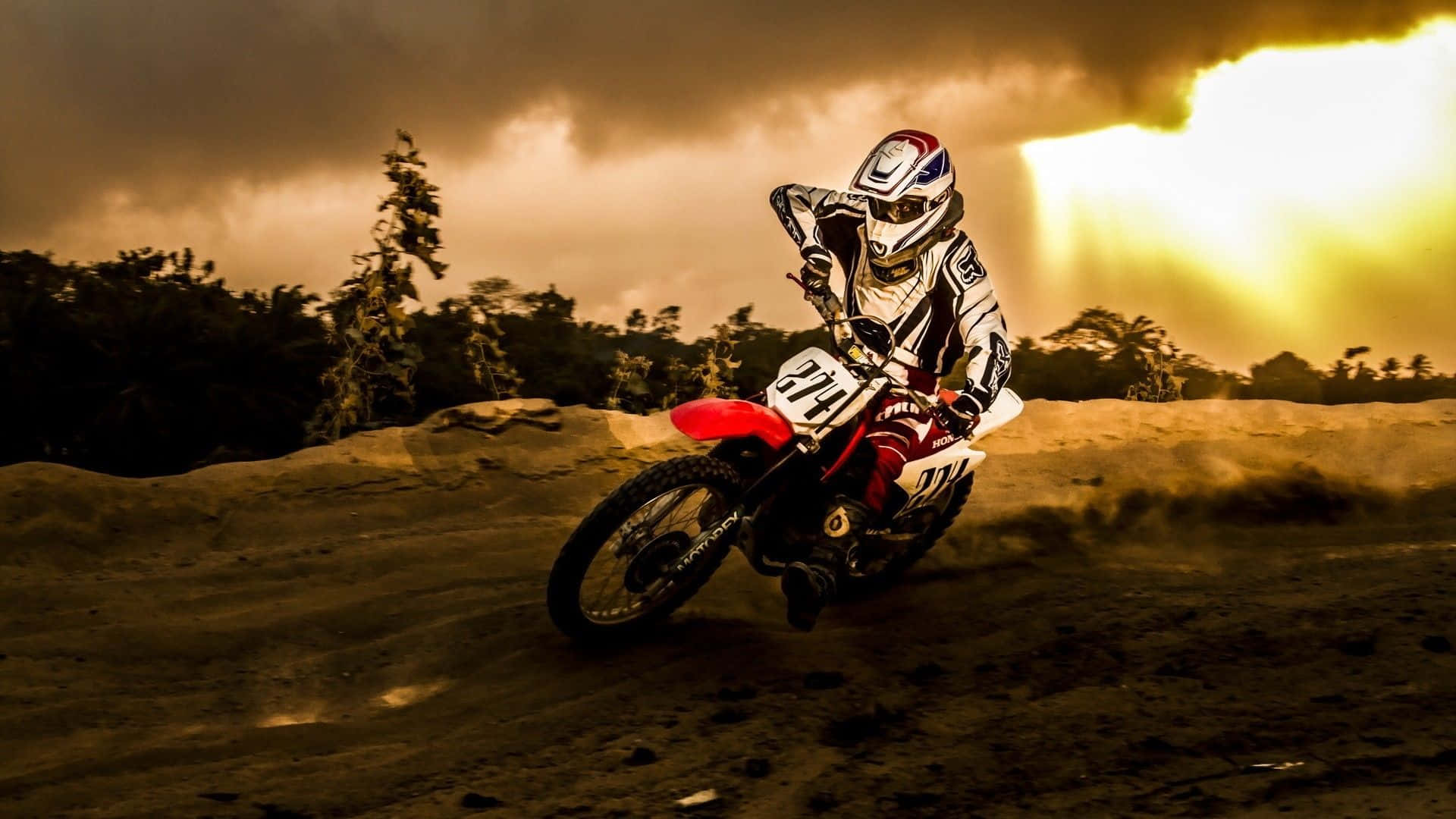 Dirtbike 桌布 壁紙 — 一位越野摩托車手在泥地賽道上傾身入彎，背後的金色陽光穿過暴風雲層灑落。