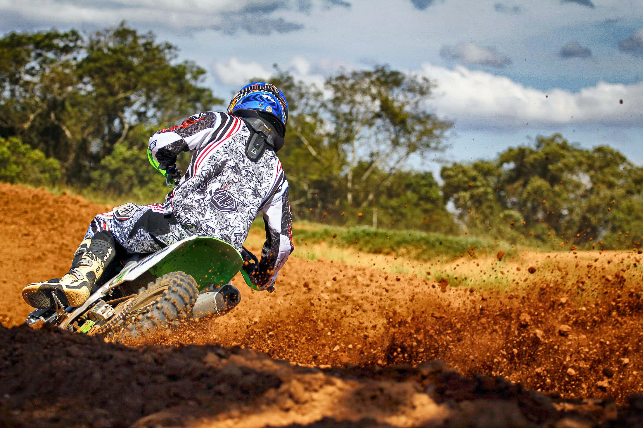 Dirtbike 桌布 壁紙 — 一位越野摩托車手在泥地賽道上傾身過急彎，在部分多雲的天空下濺起一陣紅棕色泥漿。