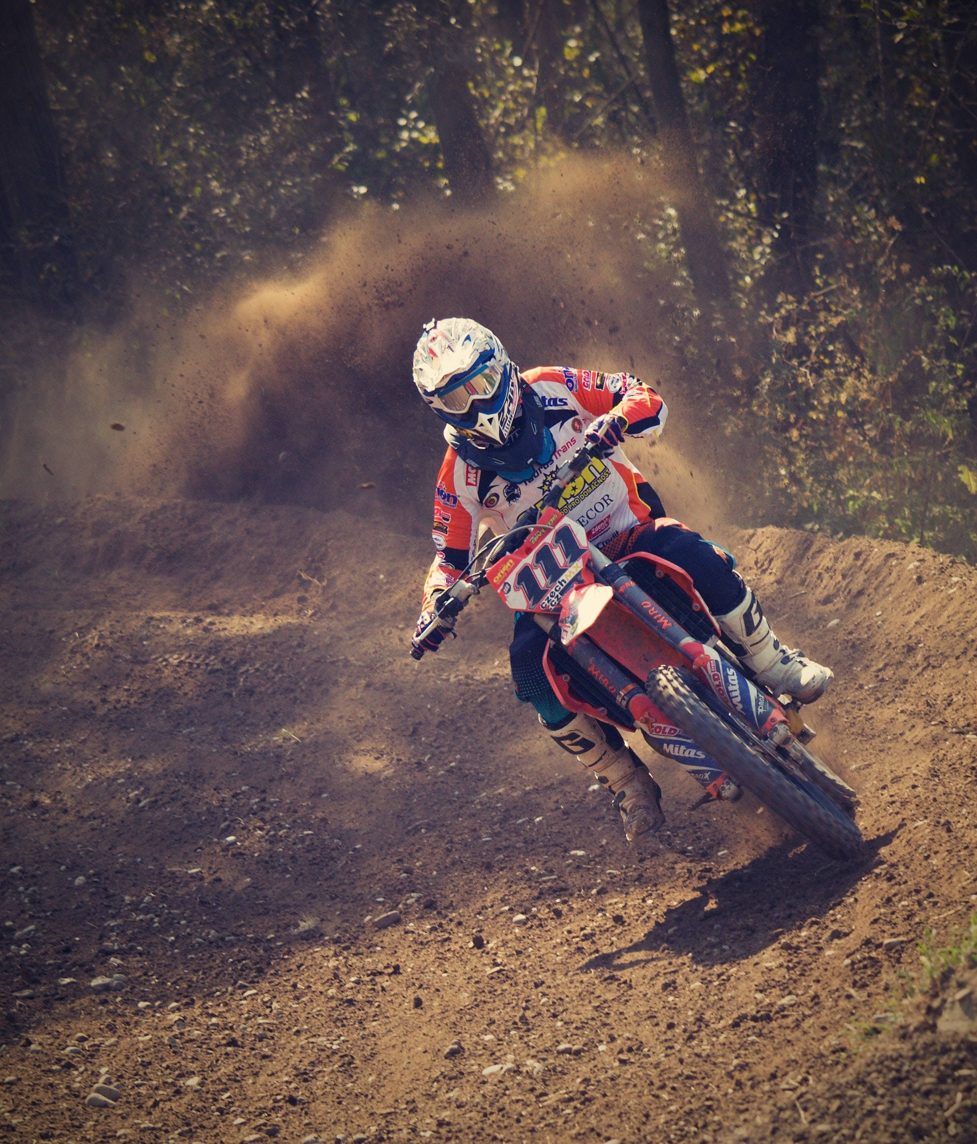 Motocross 桌布 壁紙 — 一位越野摩托車手在泥地賽道上傾身過急彎，在陽光明媚的森林中揚起一片塵土。