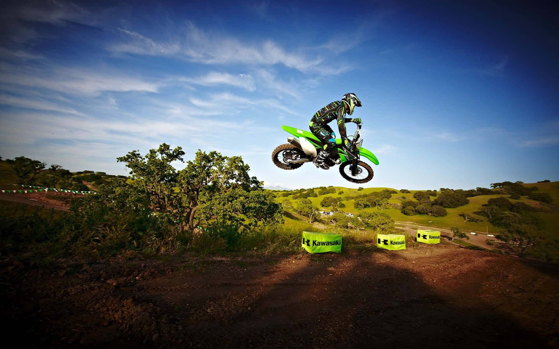 越野摩托車 桌布 壁紙 — 一位越野摩托車手在綠色 Kawasaki 摩托車上，飛越泥土賽道，下方是翠綠的山丘和藍天。