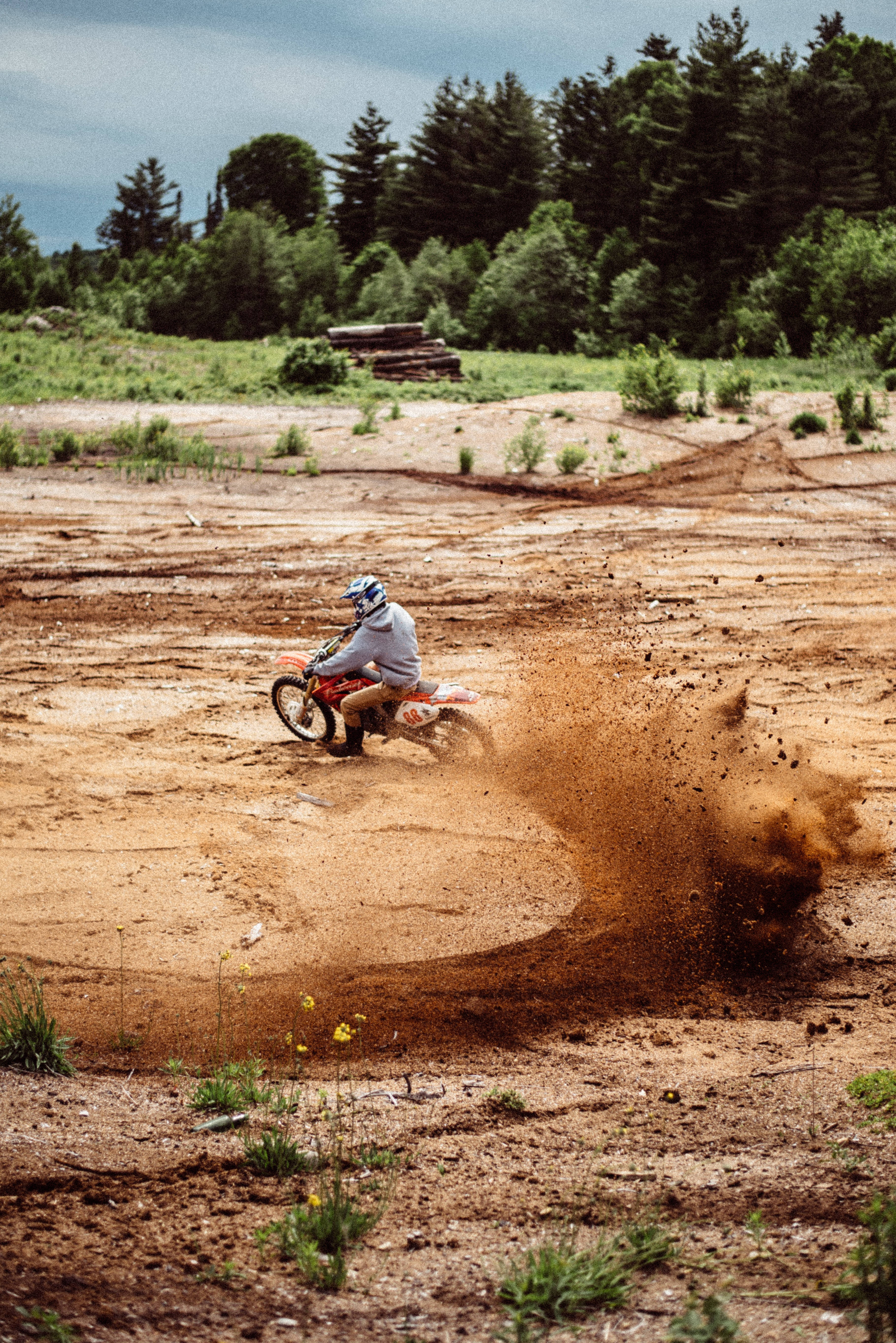 Dirtbike 桌布 壁紙 — 一位越野摩托車手在陰天之下，於塵土飛揚的泥濘賽道上飛馳，揚起一大片紅褐色的泥雲。