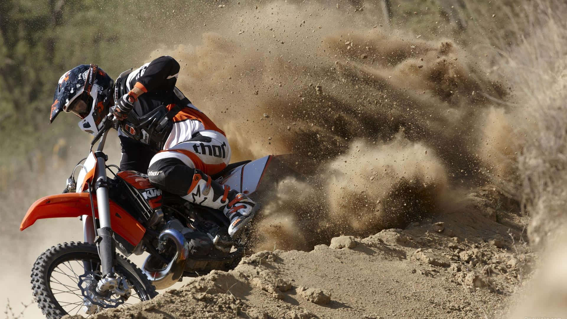 Motocross 桌布 壁紙 — 一位越野摩托車手在陽光明媚的小徑上，騎著橙色越野摩托車傾斜入急彎，揚起一大片棕色塵土。