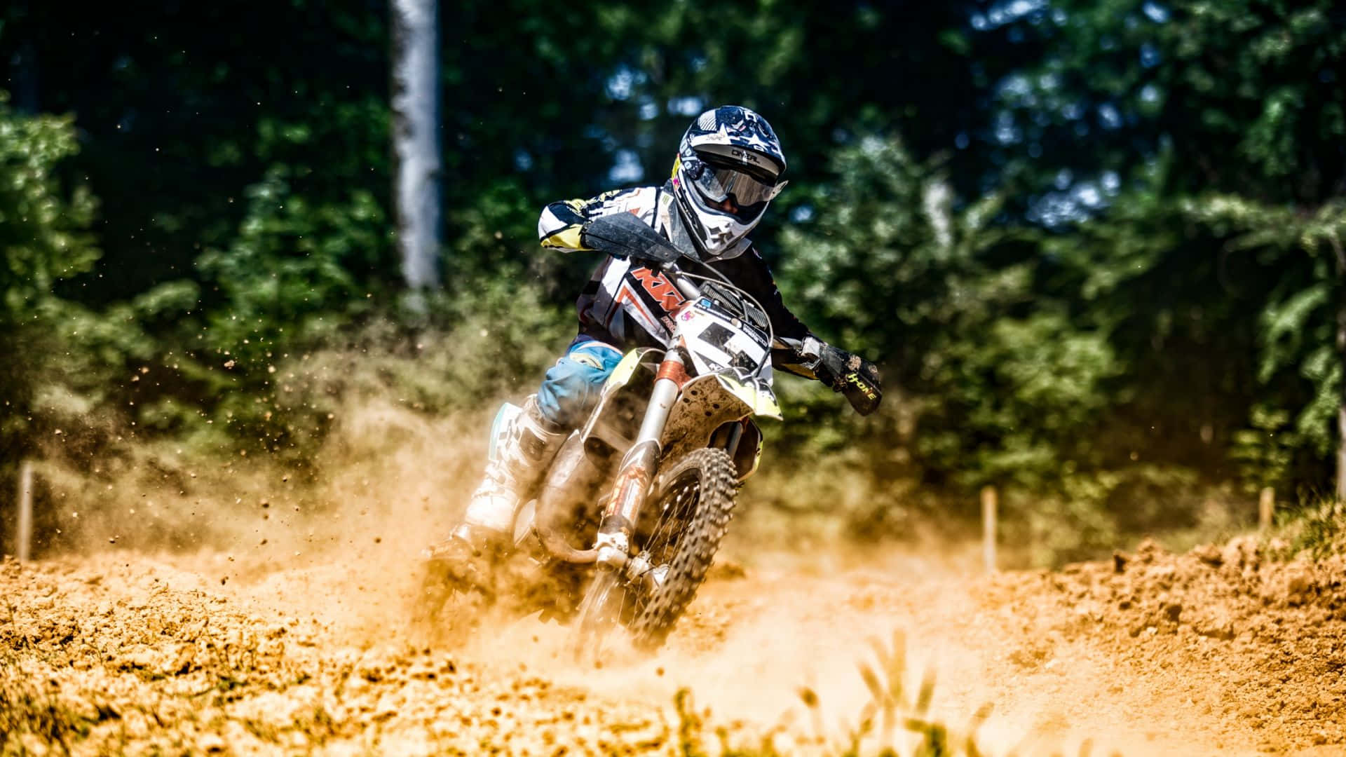Motocross 桌布 壁紙 — 一位越野摩托車手在陽光明媚的森林中的泥地賽道上傾斜入彎，揚起灰塵。