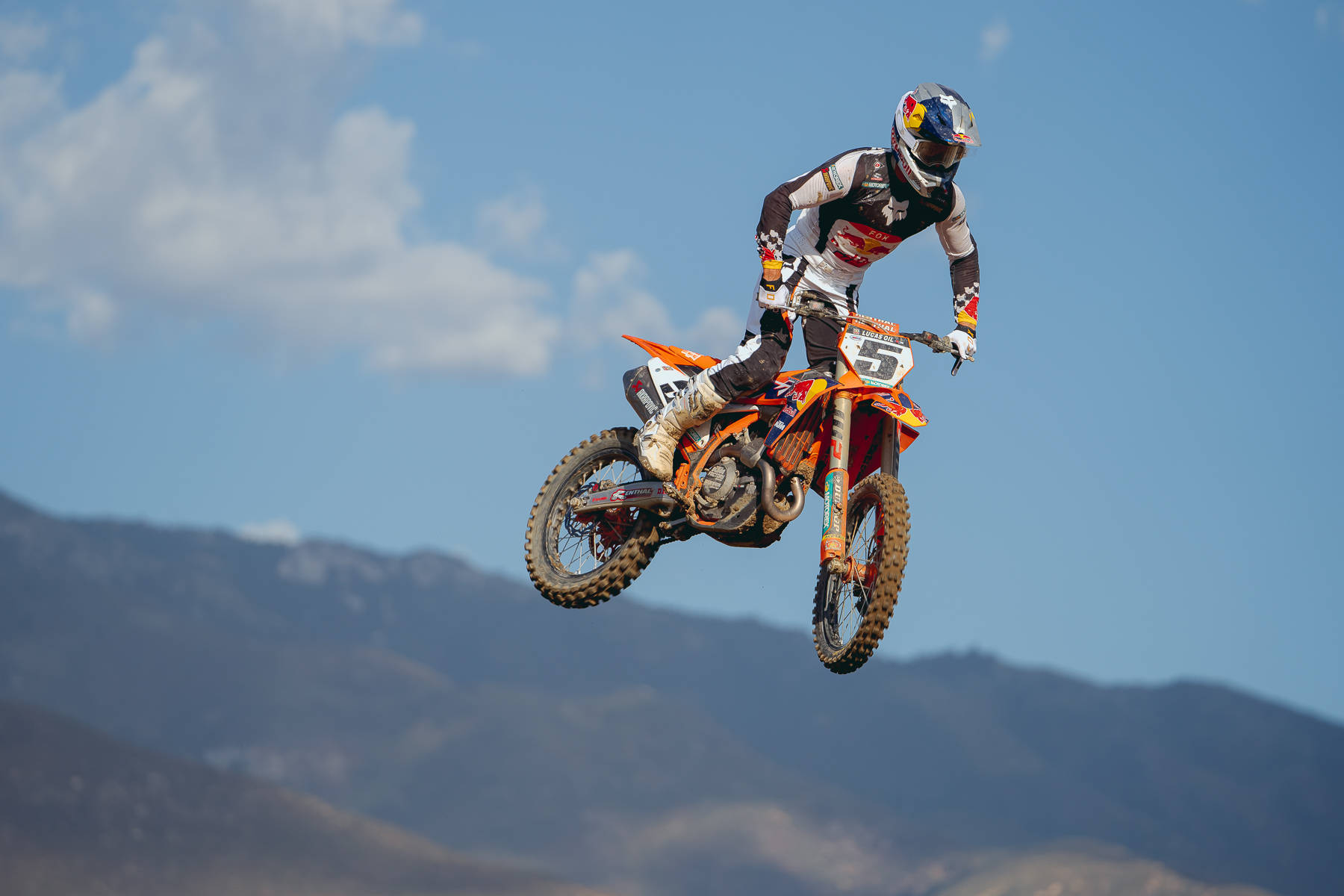 Motocross 桌布 壁紙 — 一位越野摩托車手騎著橙色摩托車在空中飛翔，背景是遠山和晴朗的藍天。