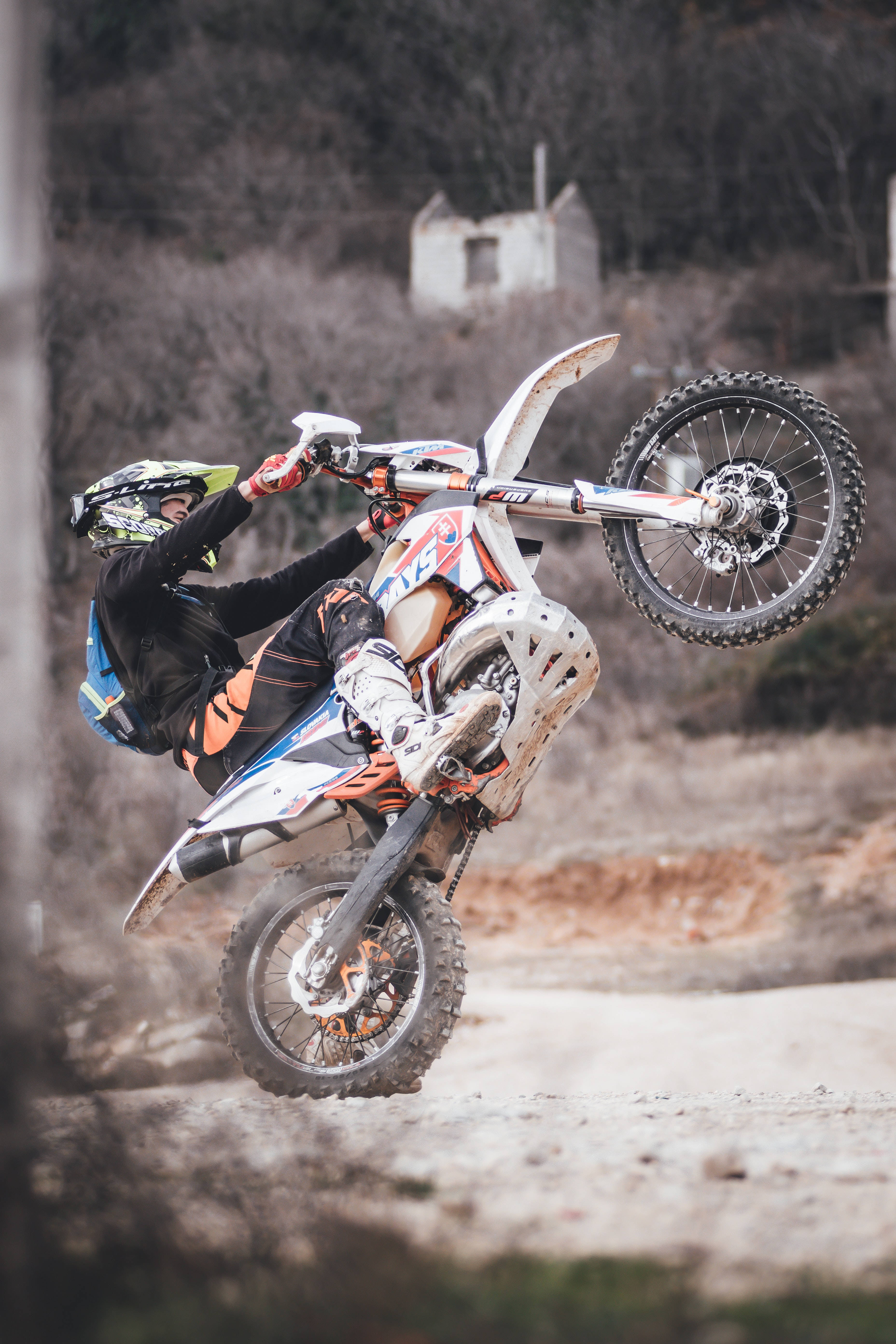 Dirtbike 桌布 壁紙 — 一位越野機車手在崎嶇的泥路上大膽表演翹前輪，背景是一座破舊的建築物。