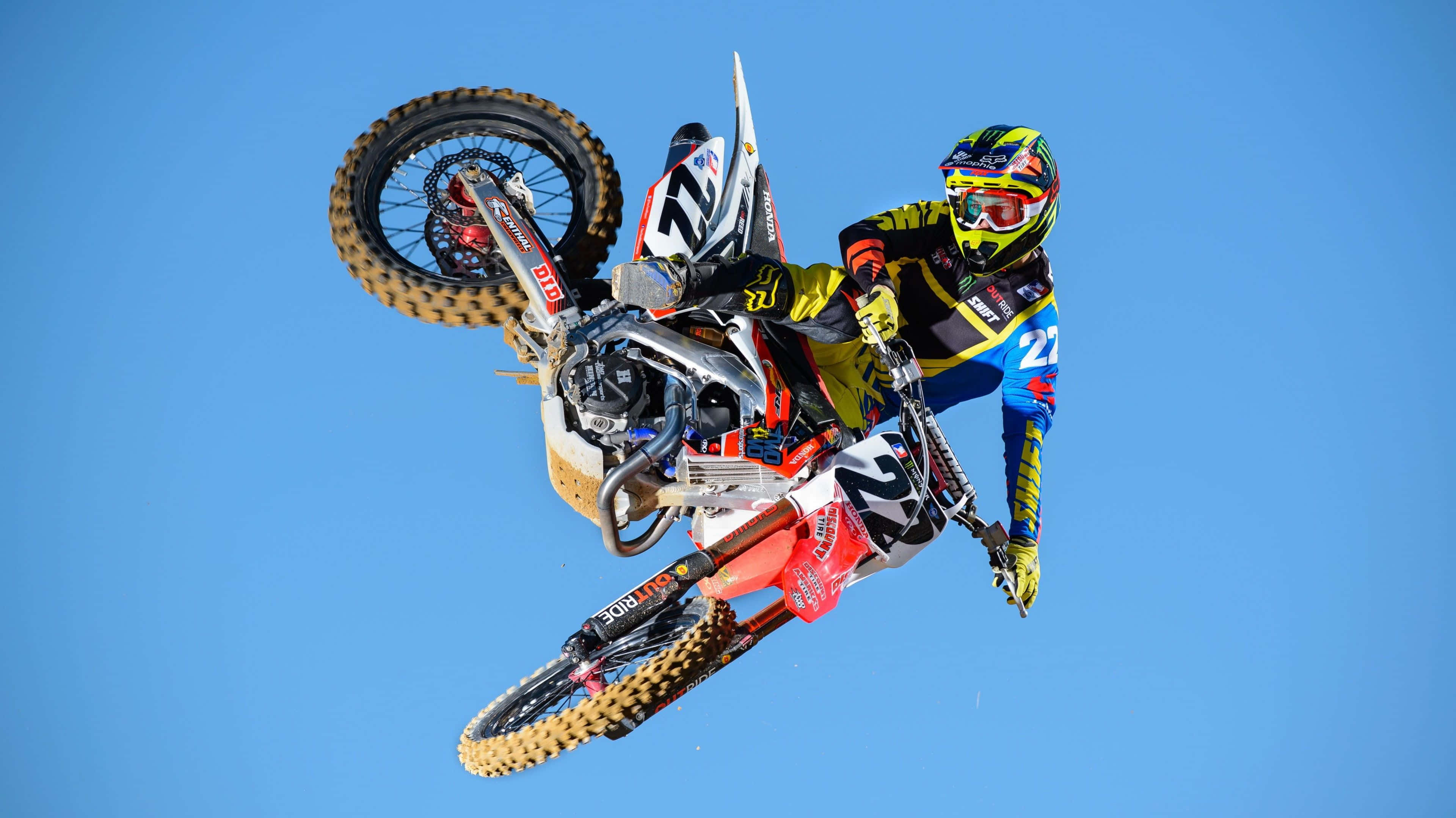 Motocross 桌布 壁紙 — 一位越野機車車手在晴朗的藍天中飛翔，在越野機車上表演大膽的後空翻特技。