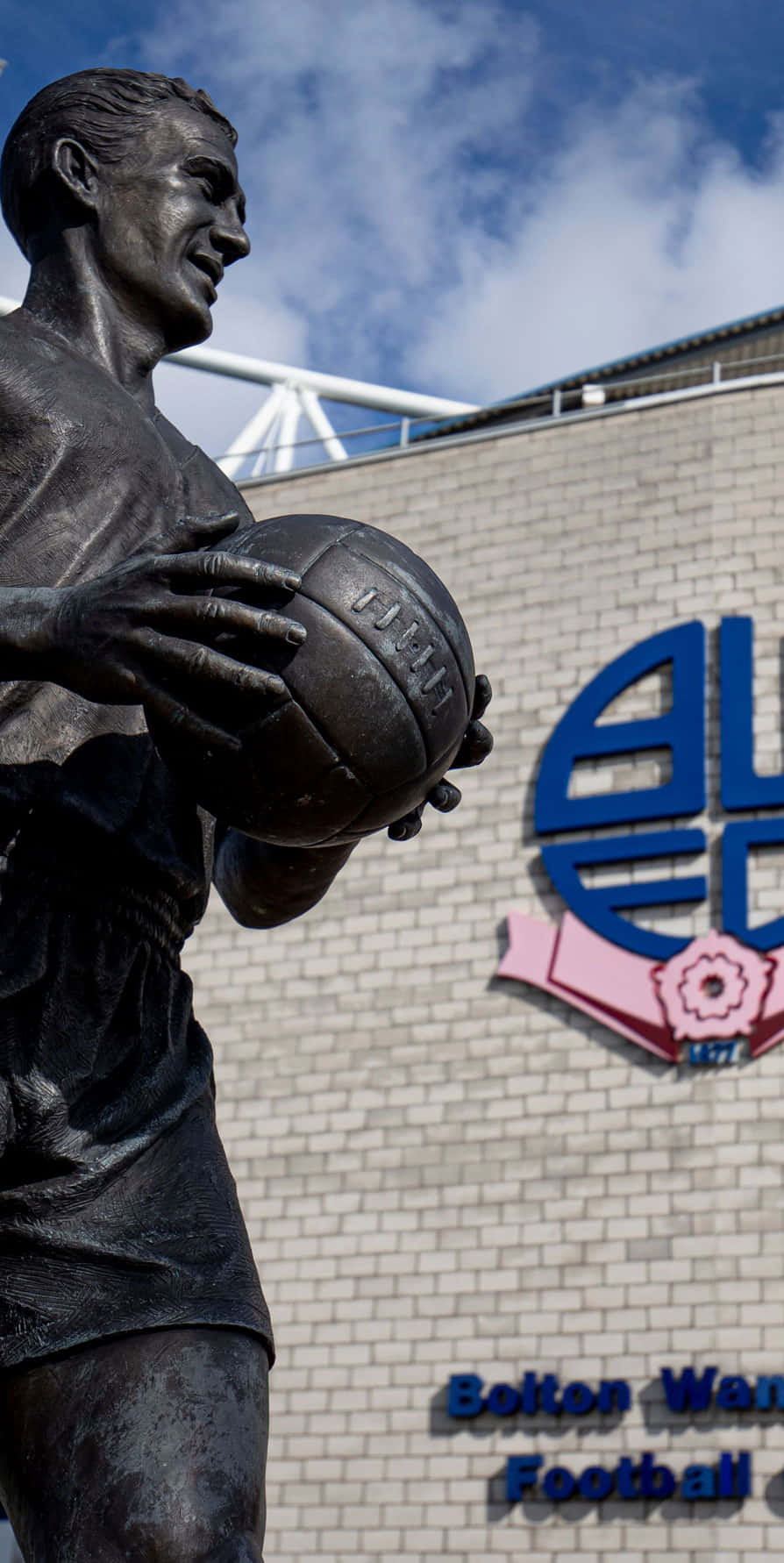Bolton Wanderers Fc 桌布 壁紙 — 一位足球員手持足球的銅像，在部分多雲的天空下，自豪地矗立在帶有博爾頓流浪者隊徽的磚牆前。