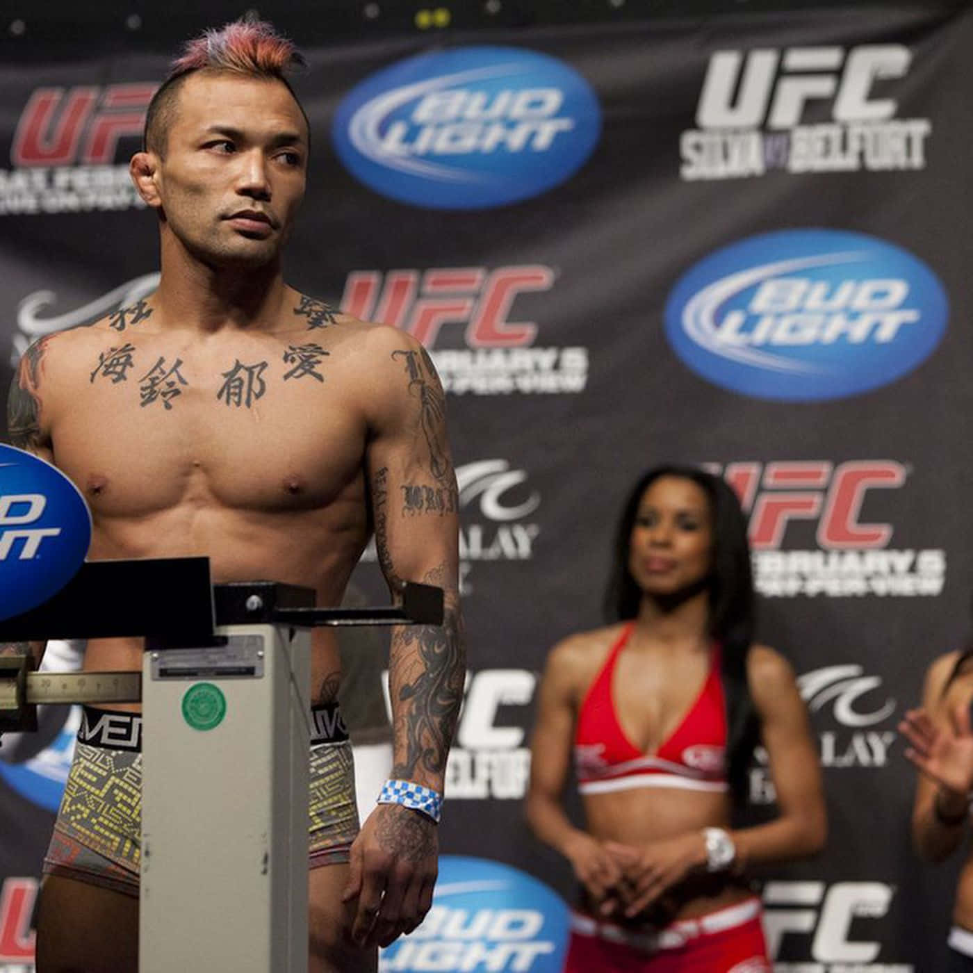 UFC 桌布 壁紙 — 一位身上有刺青、留著粉紅色莫西干頭的UFC拳手在稱重活動中站在體重計上，背景中有一位身穿比基尼的擂台女郎。