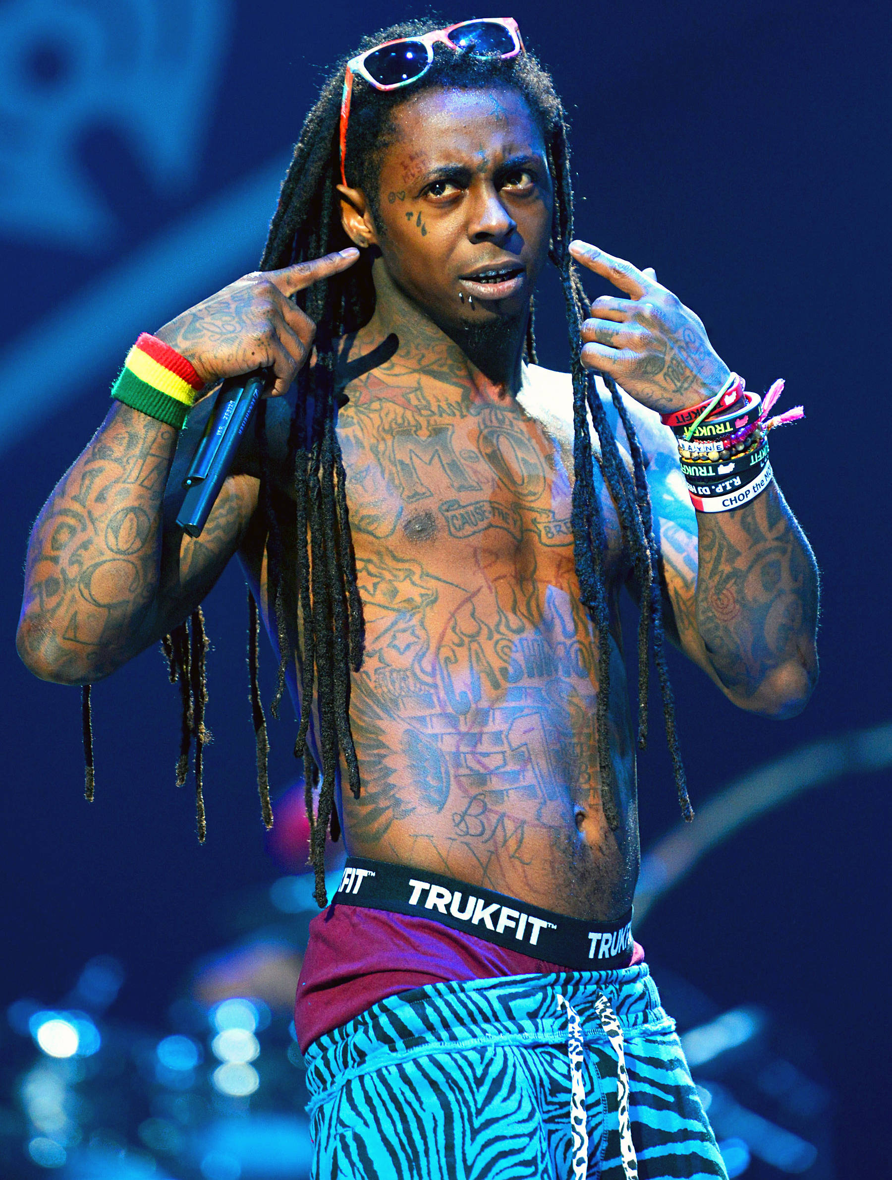 Lil Wayne 桌布 壁紙 — 一位身上有刺青、頭戴太陽眼鏡、留著雷鬼辮的音樂家在昏暗的舞台上指著自己的太陽穴。