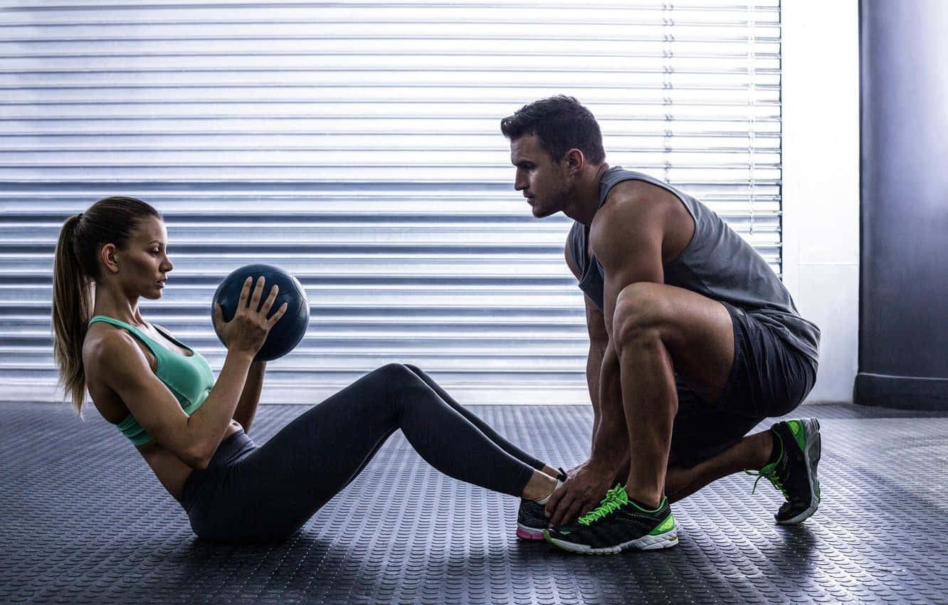 Exercise 桌布 壁紙 — 一位身材健美的女性拿著藥球，而一位男性以深蹲姿勢在金屬地板和百葉窗的健身房中協助她。
