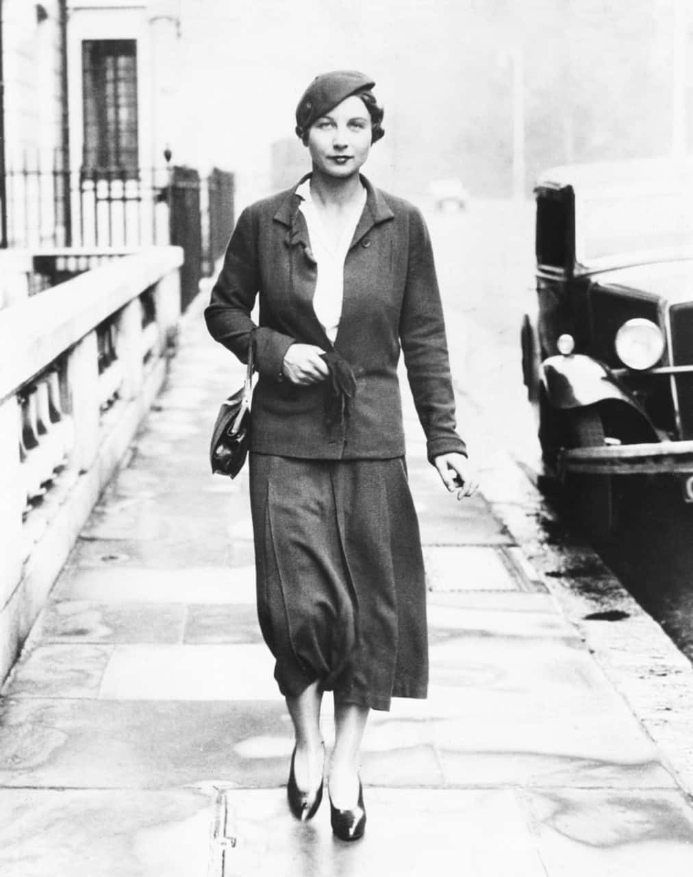 Tennis 桌布 壁紙 — 一位身穿1930年代西裝和貝雷帽的女性，自信地走在濕漉漉的城市人行道上，旁邊是一輛老爺車。