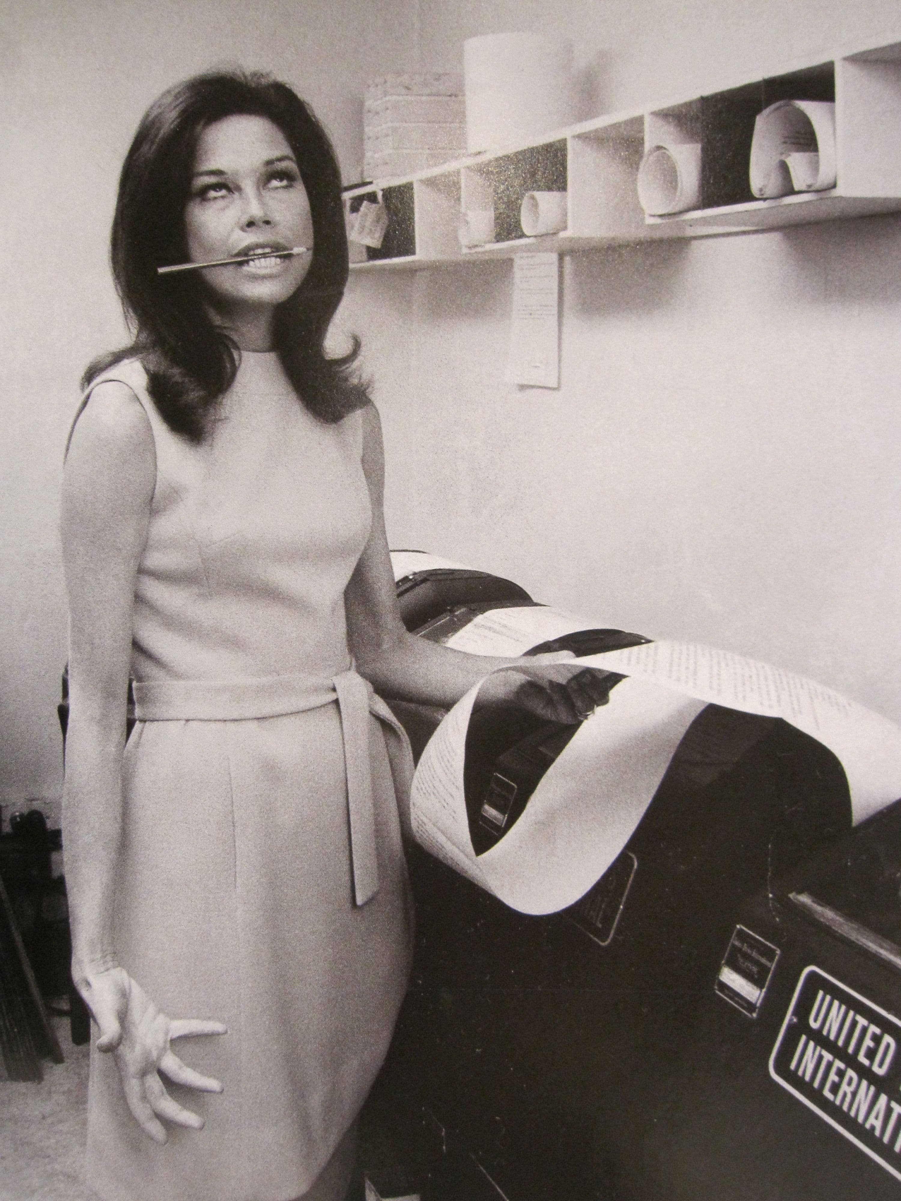 復古 桌布 壁紙 — 一位身穿1960年代連衣裙的女性，嘴裡叼著一支筆，站在一台United International郵件機旁邊，身處單色調的辦公室環境中。
