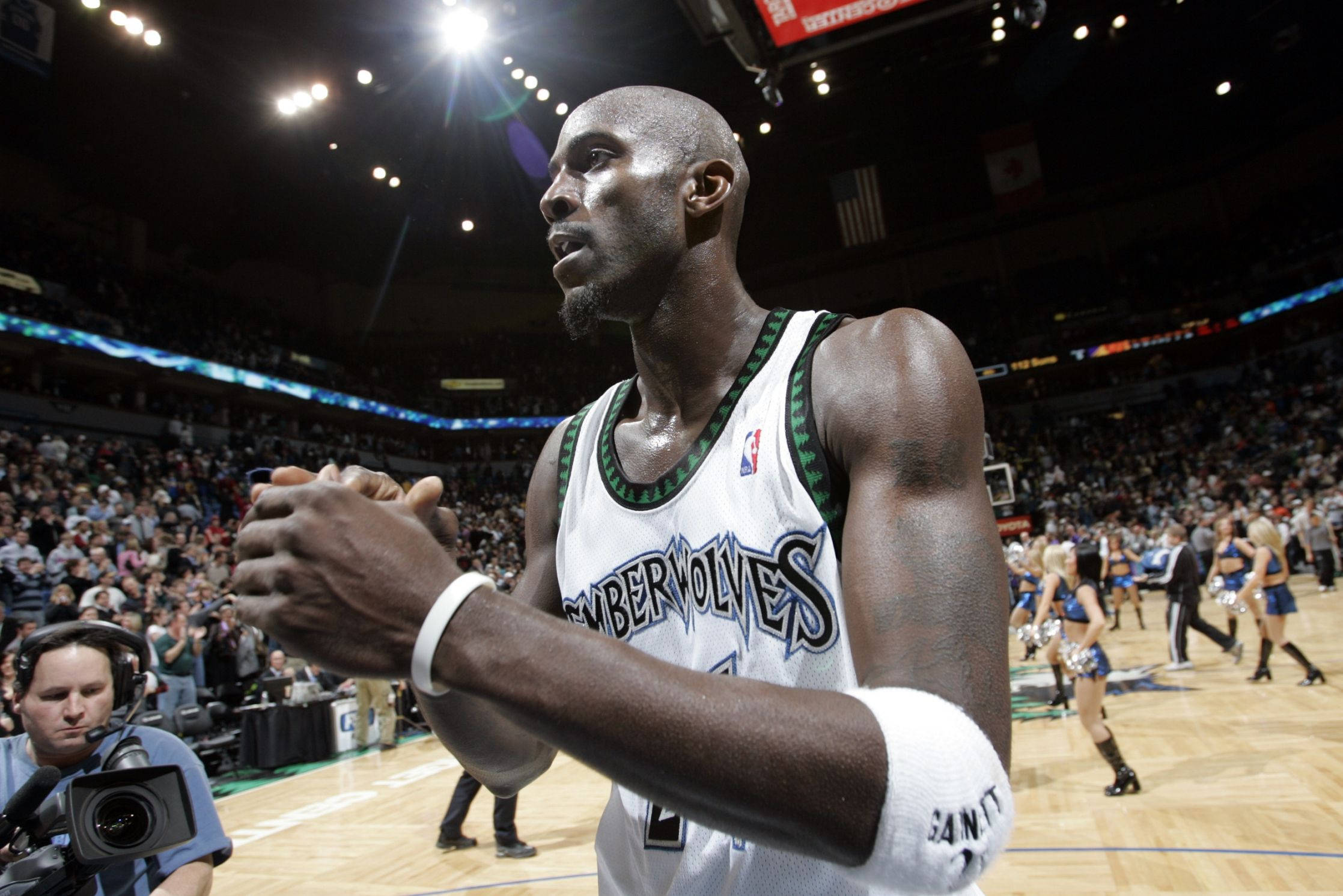 Kevin Garnett 桌布 壁紙 — 一位身穿Timberwolves球衣的專注籃球運動員站在燈光明亮的球場上，背景有群眾和啦啦隊。