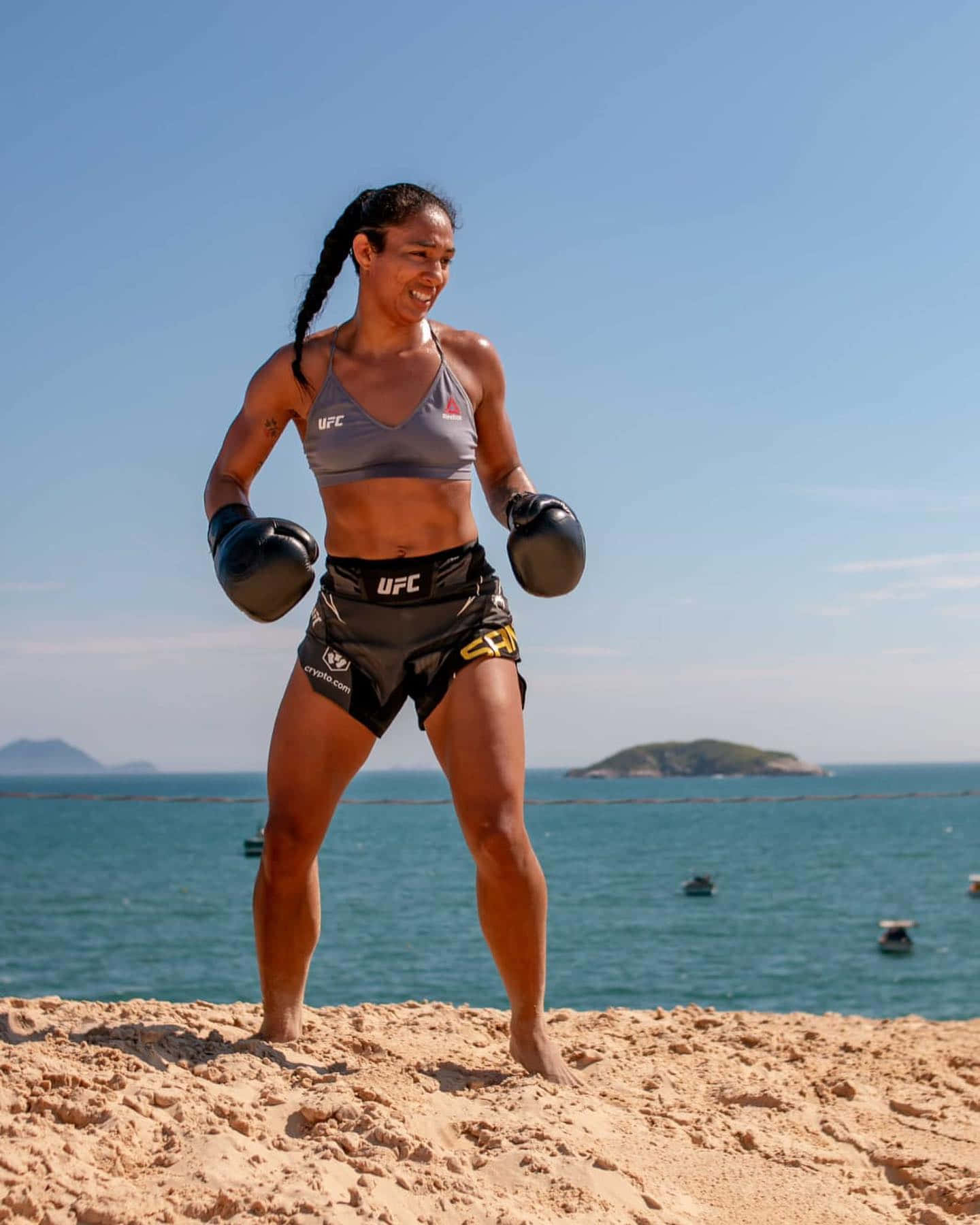 Fitness 桌布 壁紙 — 一位身穿UFC裝備的女性MMA拳手在陽光明媚的海灘上自信微笑，背景是碧綠的海洋和遠處的島嶼。