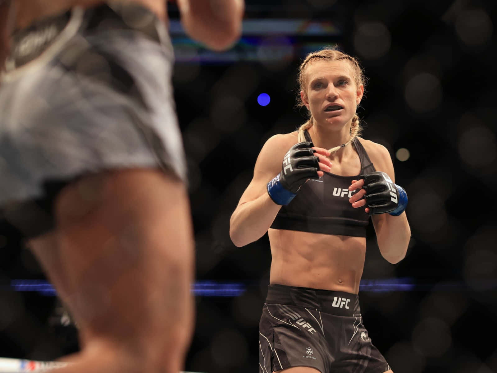 桌布 壁紙 — 一位身穿UFC裝備的女性MMA拳手在八角籠中專注站立，而她的對手則在前景中隱約出現。