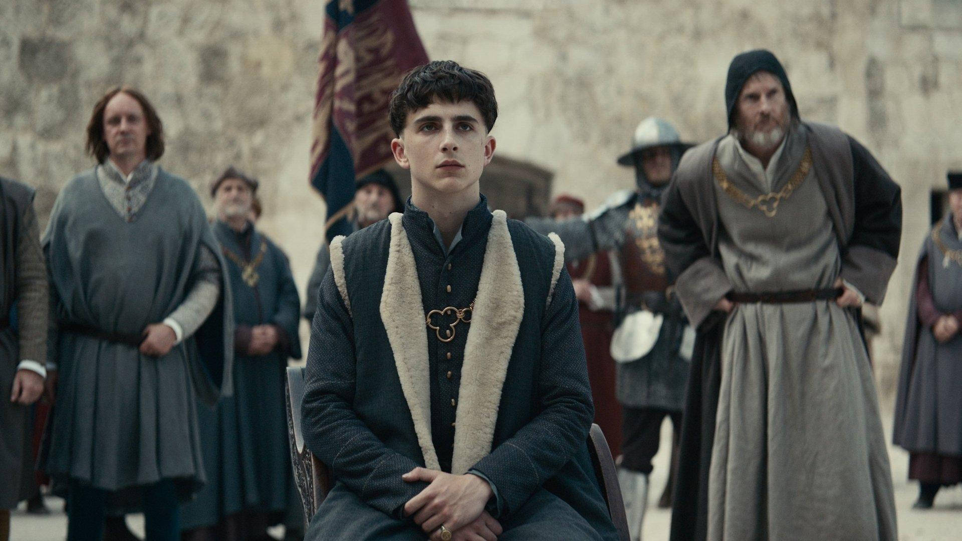 Timothée Chalamet 桌布 壁紙 — 一位身穿中世紀服飾的年輕男子莊嚴地坐在石牆前，身後站著穿著時代服裝的人物。