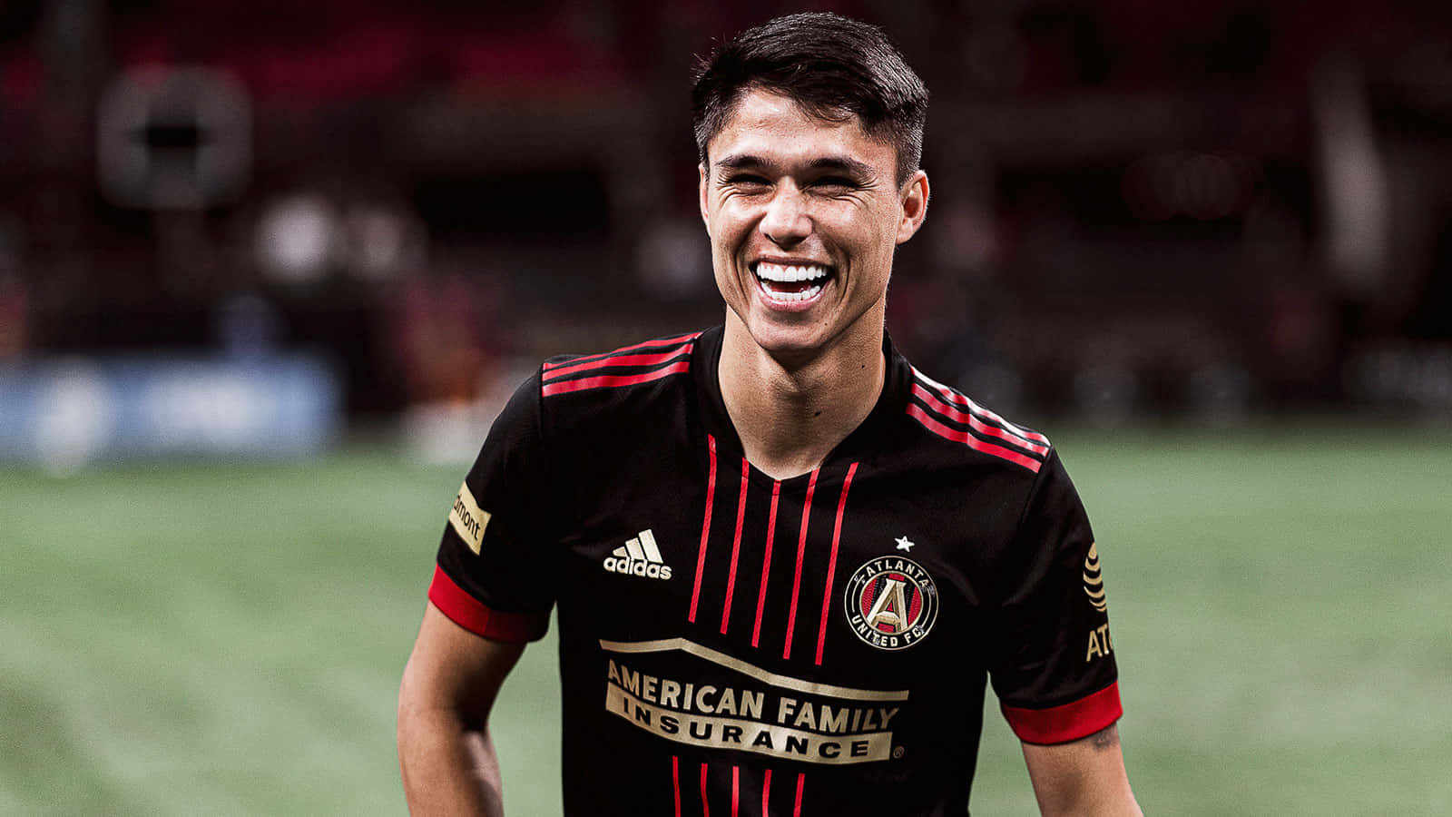 Athletic 桌布 壁紙 — 一位身穿亞特蘭大聯隊黑紅球衣的快樂足球運動員在球場燈光下的綠色草地上開懷大笑。