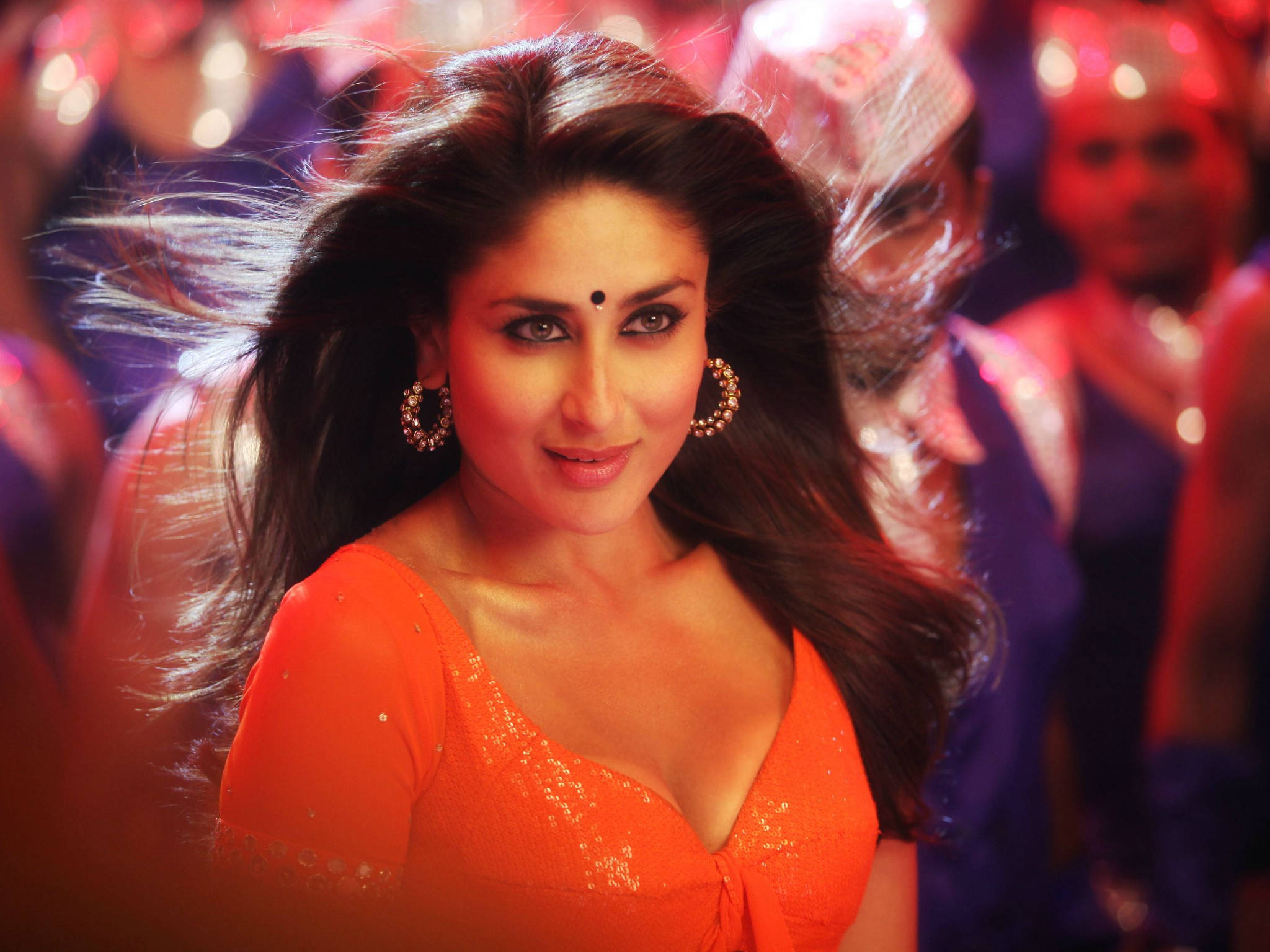 Kareena 桌布 壁紙 — 一位身穿亮橙色亮片連衣裙的女性自信地微笑，她的深色頭髮在模糊的節日背景下飄動。