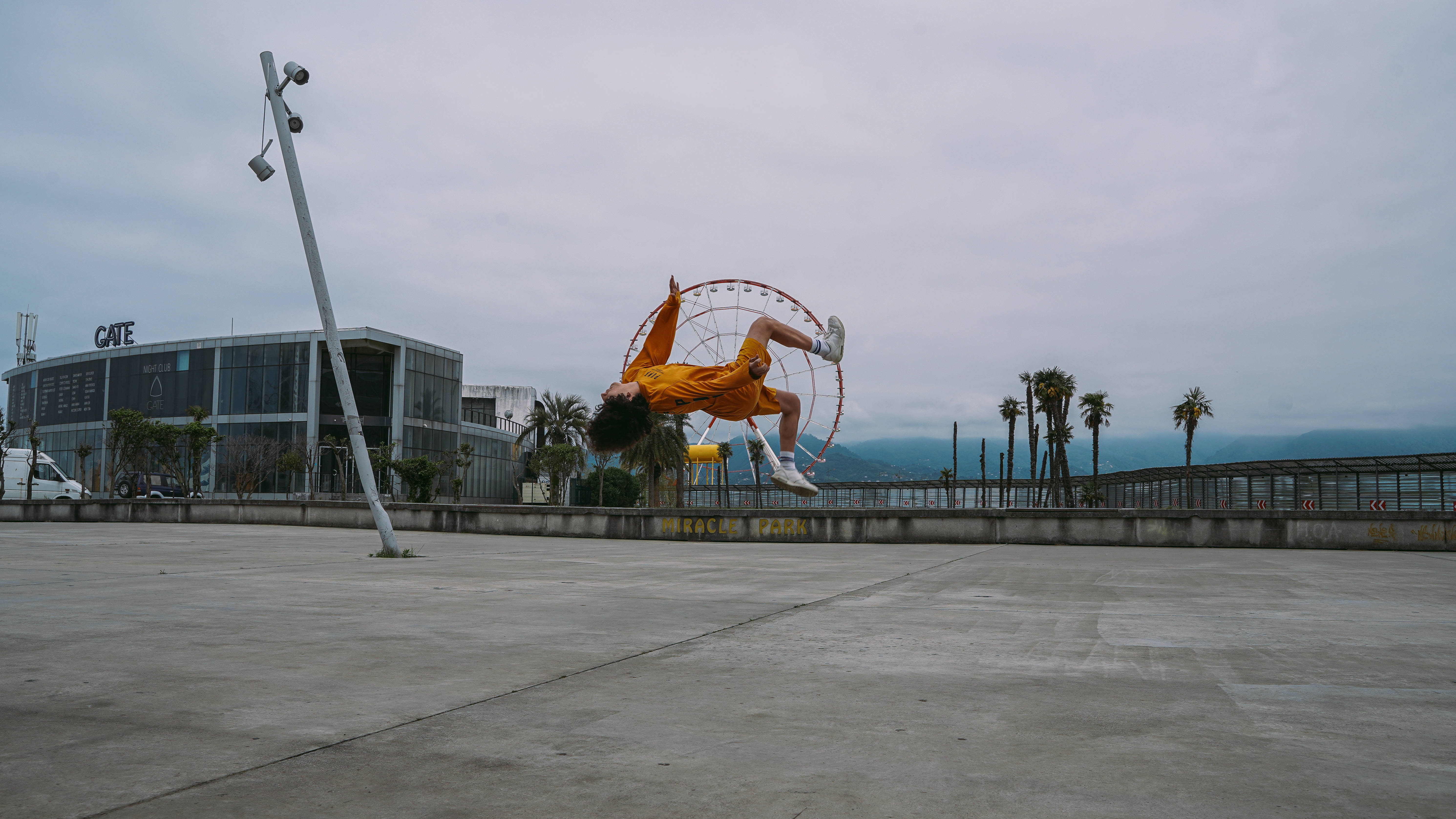 Parkour 桌布 壁紙 — 一位身穿亮橙色服裝的霹靂舞者在陰天下的寬敞城市廣場中的圓形框架上表演空中旋轉。