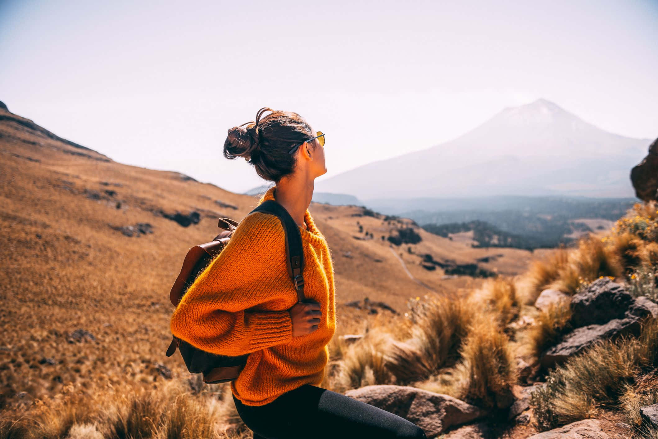 Travel Desktop 桌布 壁紙 — 一位身穿亮橙色毛衣的女性坐在岩石山坡上，凝視著晴朗天空下遠處的雪山。