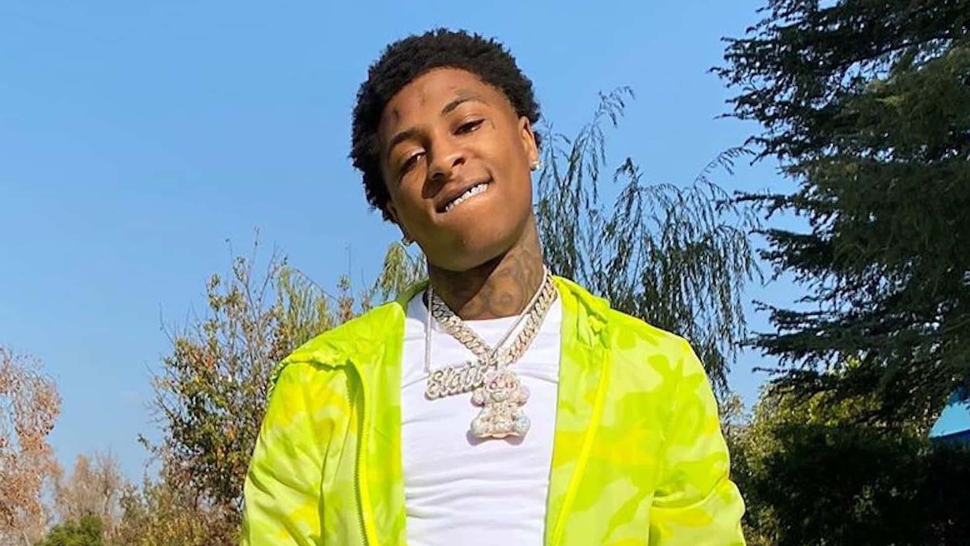 Youngboy 桌布 壁紙 — 一位身穿亮眼螢光黃外套的年輕男子，在晴朗藍天和身後綠樹的襯托下自信地微笑。