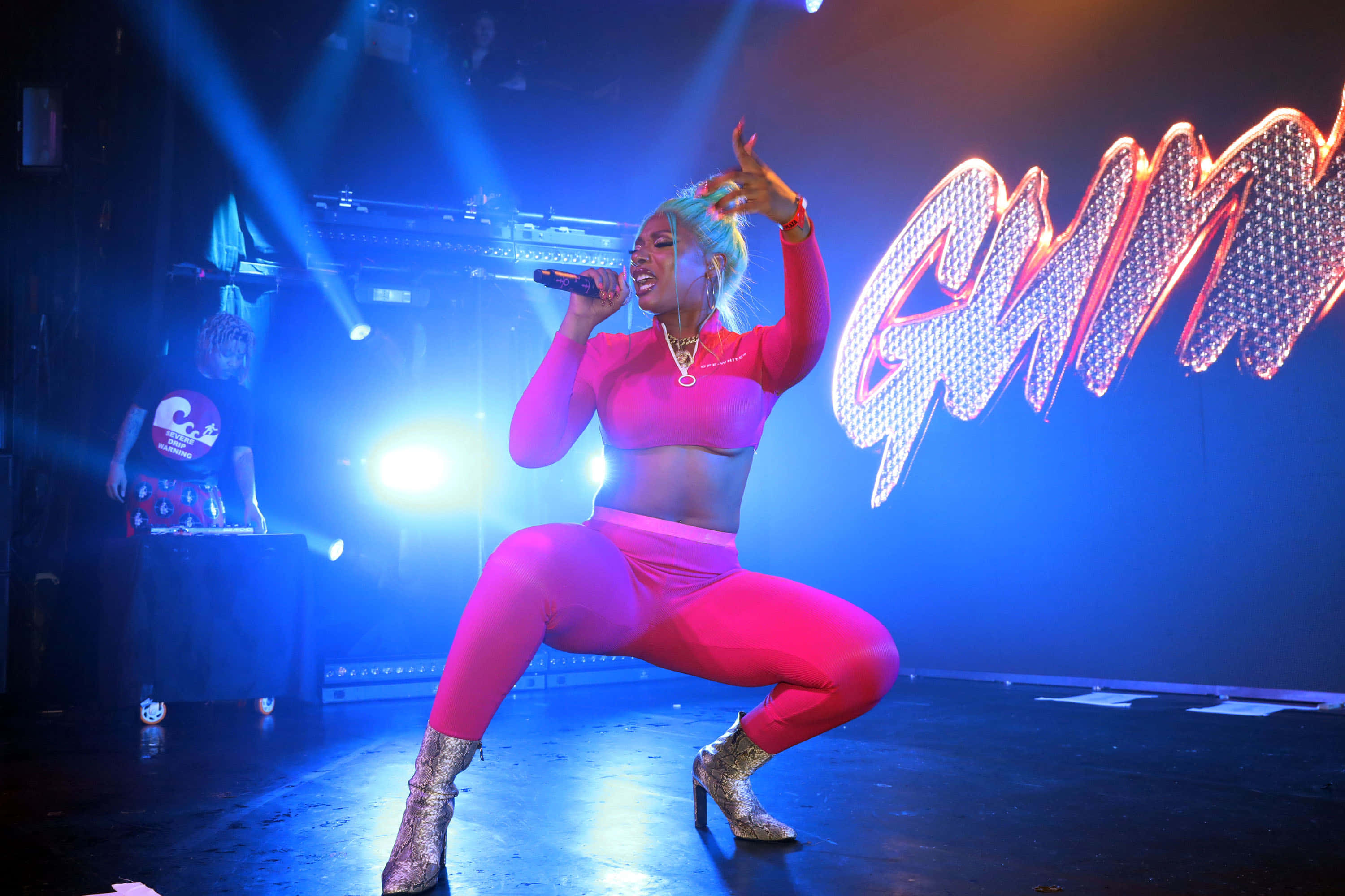 Megan Thee Stallion 桌布 壁紙 — 一位身穿亮粉紅色服裝的女表演者在藍色燈光的舞台上熱情歌唱，身後有閃爍的「GUMMY」標誌。