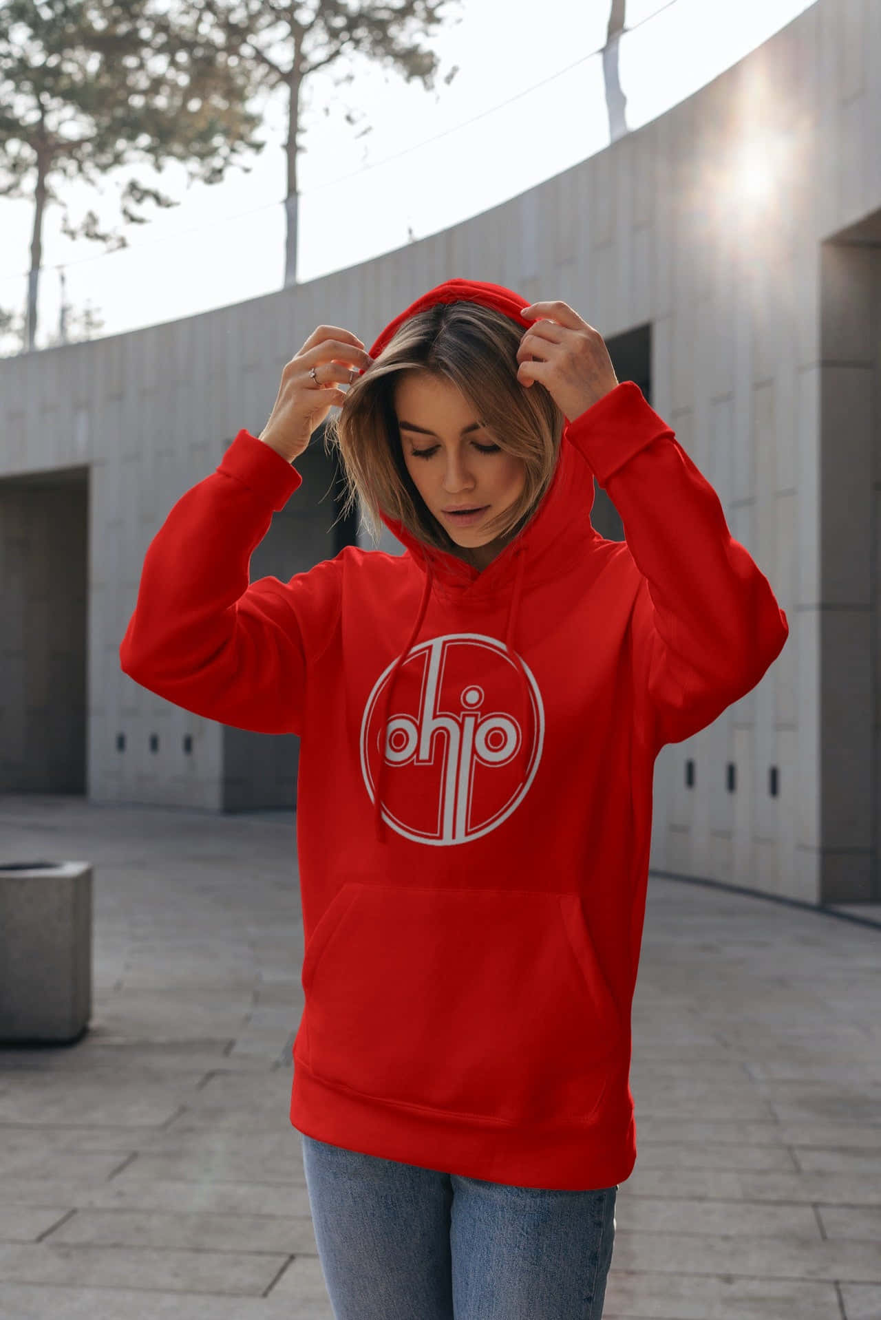 Red Hoodie 桌布 壁紙 — 一位身穿亮紅色連帽衫的女性在現代都市庭院中調整帽子，陽光穿過樹木灑落下來。