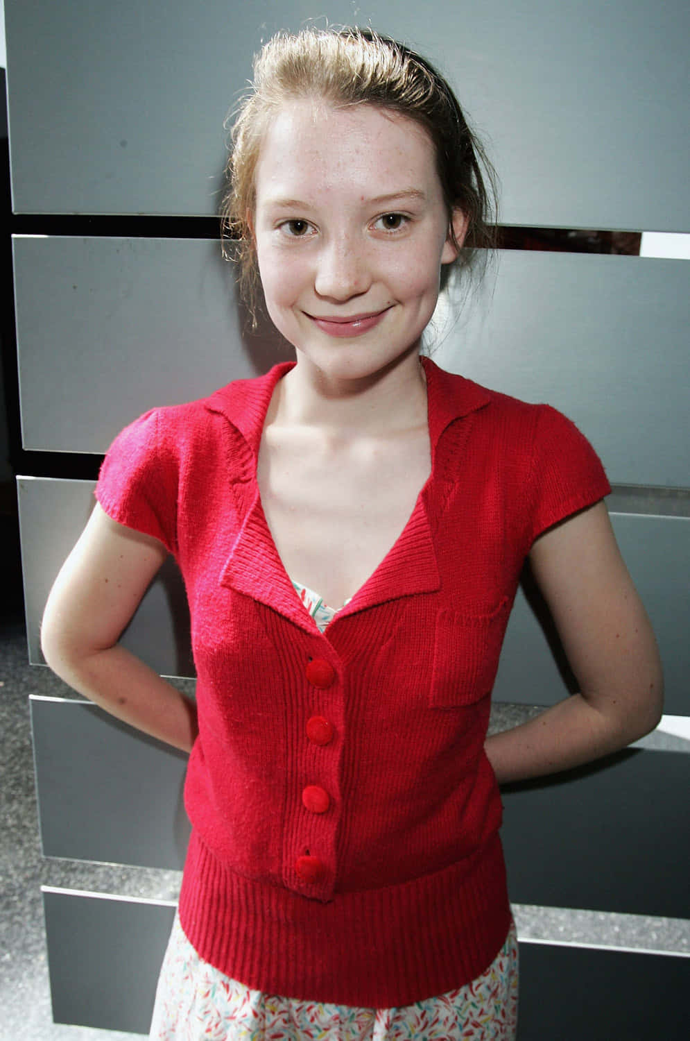Mia Wasikowska 桌布 壁紙 — 一位身穿亮紅色針織開衫的年輕女孩，在現代灰色鑲板牆前溫柔地微笑。