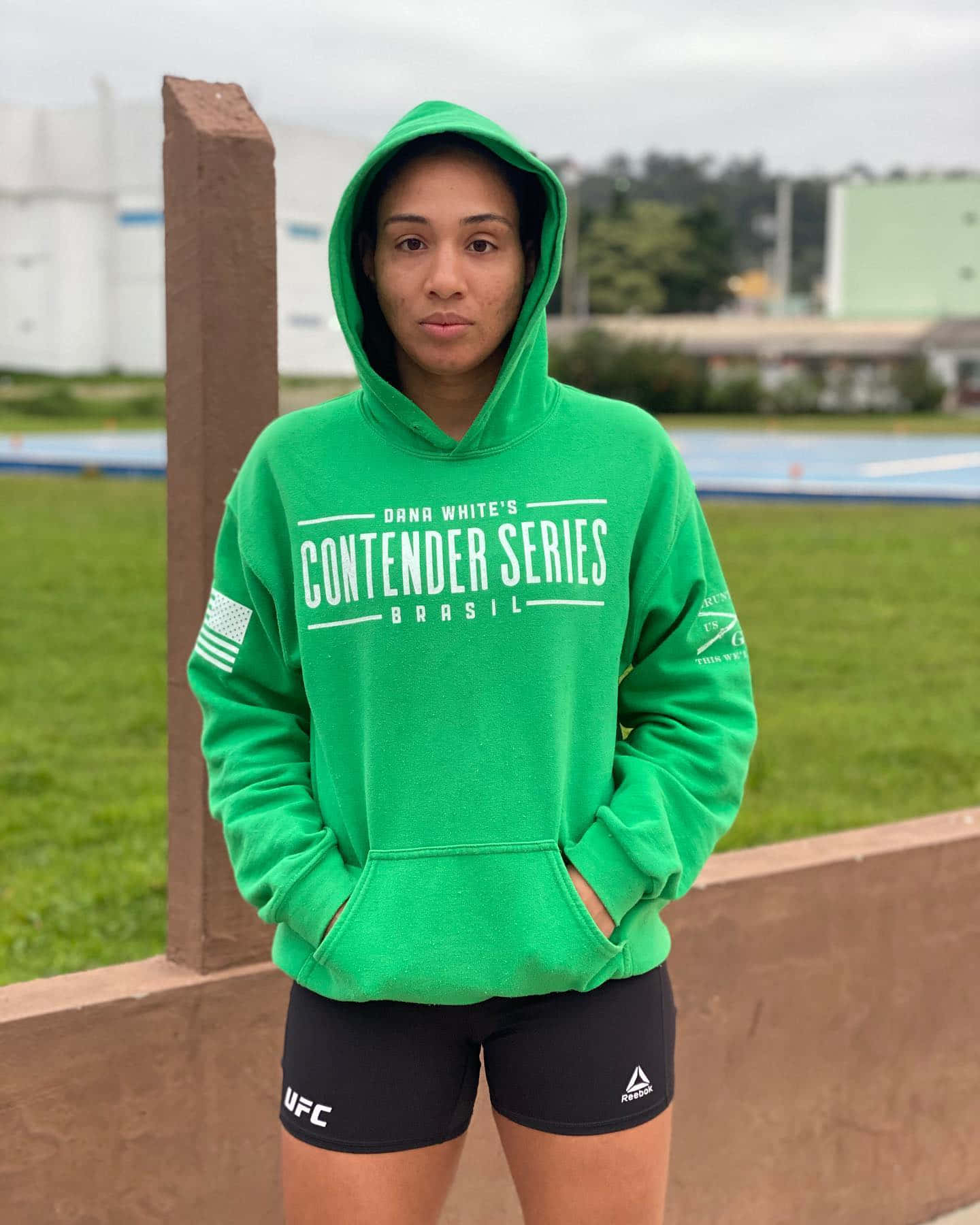 MMA 桌布 壁紙 — 一位身穿亮綠色連帽衫和黑色UFC短褲的女性，雙手插袋，自信地站在戶外跑道附近。