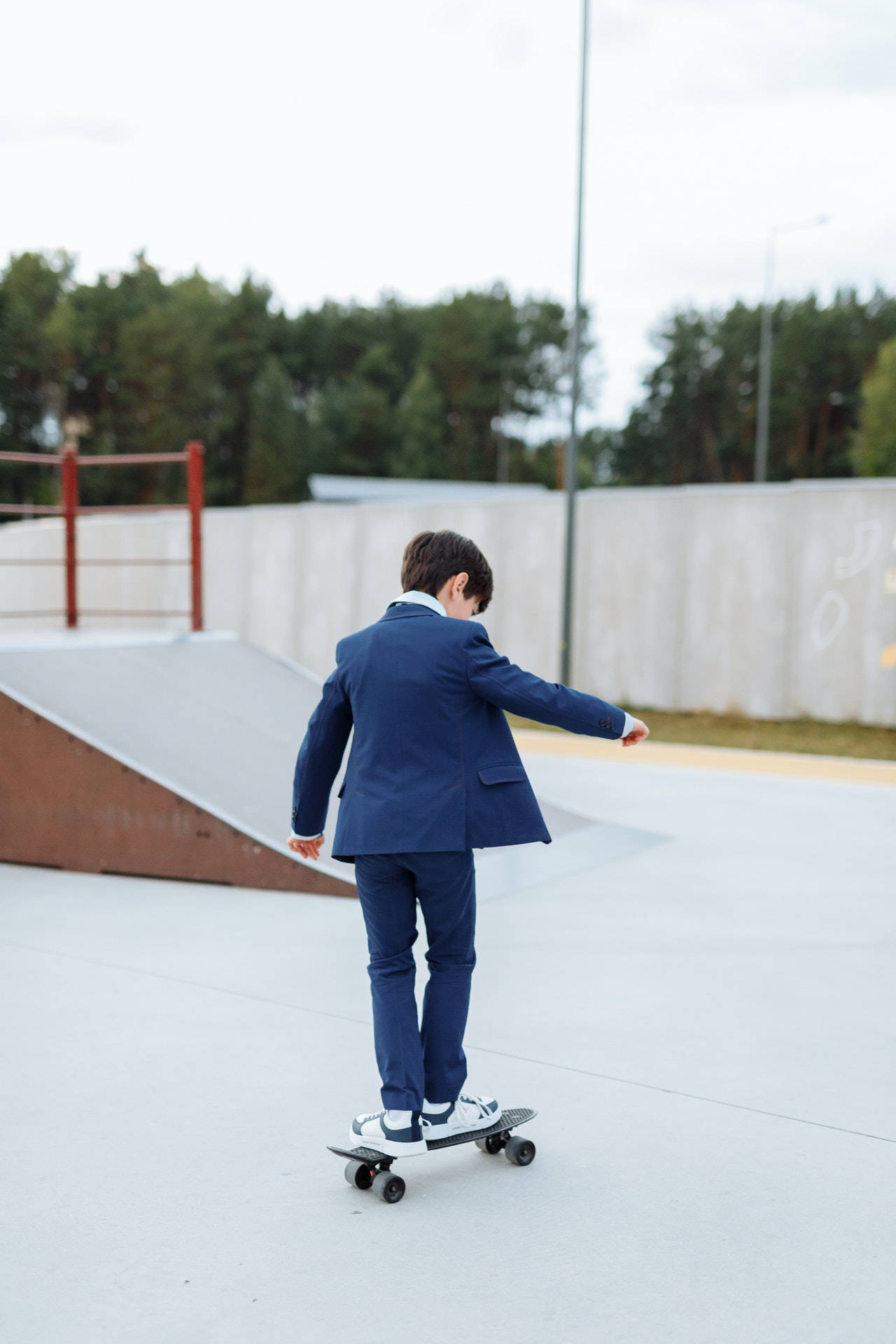 Skater Boy 桌布 壁紙 — 一位身穿亮藍色西裝的年輕男子在背景有樹木的混凝土滑板公園玩滑板。