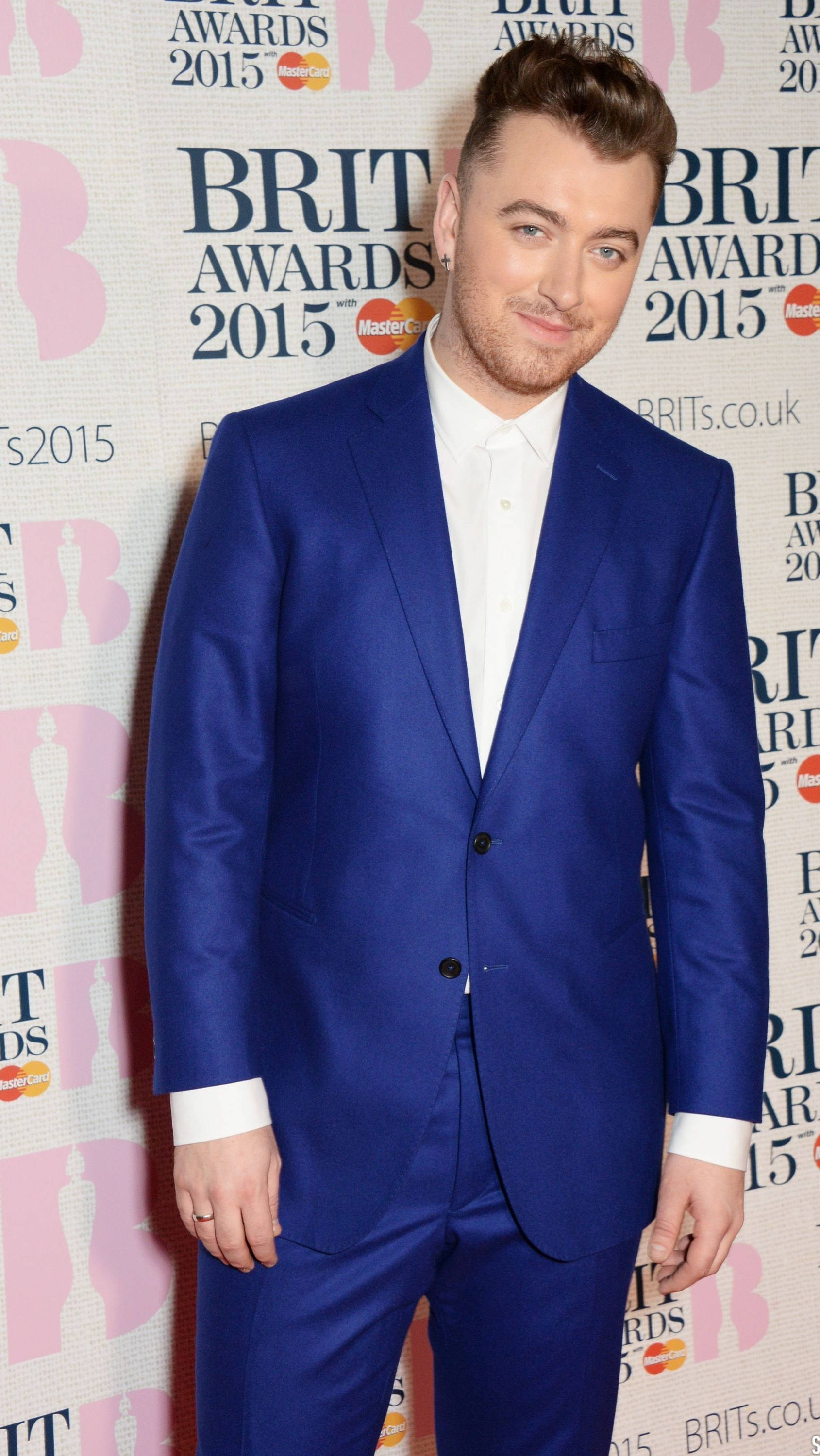Sam Smith 桌布 壁紙 — 一位身穿亮藍色西裝的男士在2015年全英音樂獎的紅地毯上自信地微笑，背景為粉紅色和白色的品牌背景。