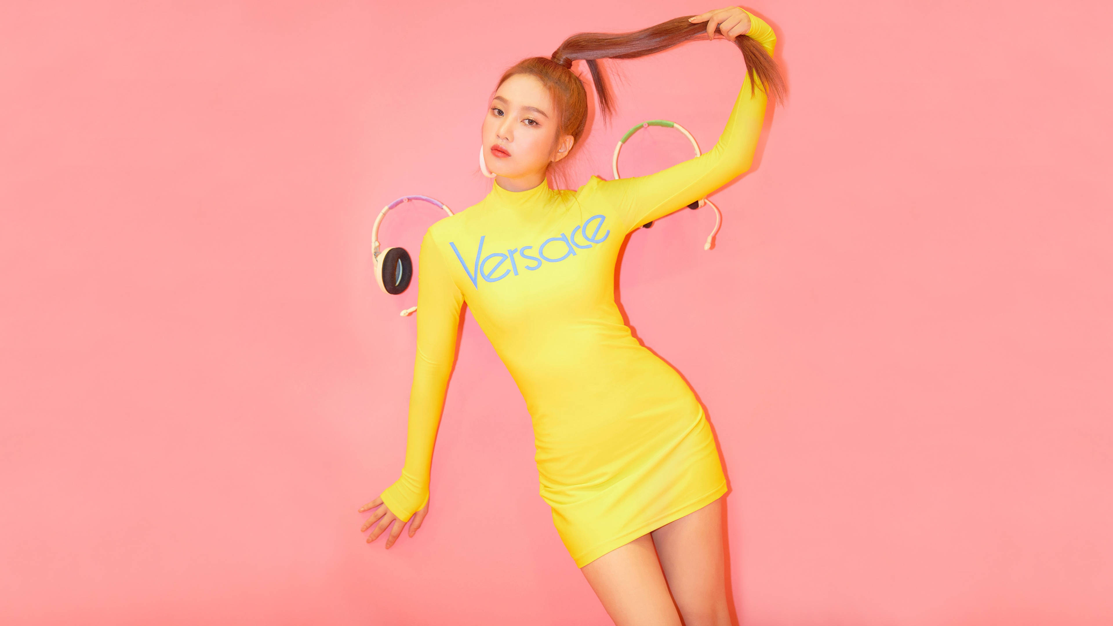 Velvet 桌布 壁紙 — 一位身穿亮黃色 Versace 連衣裙的女性在純粉色背景前擺姿勢，同時抓住馬尾辮並戴著耳機。