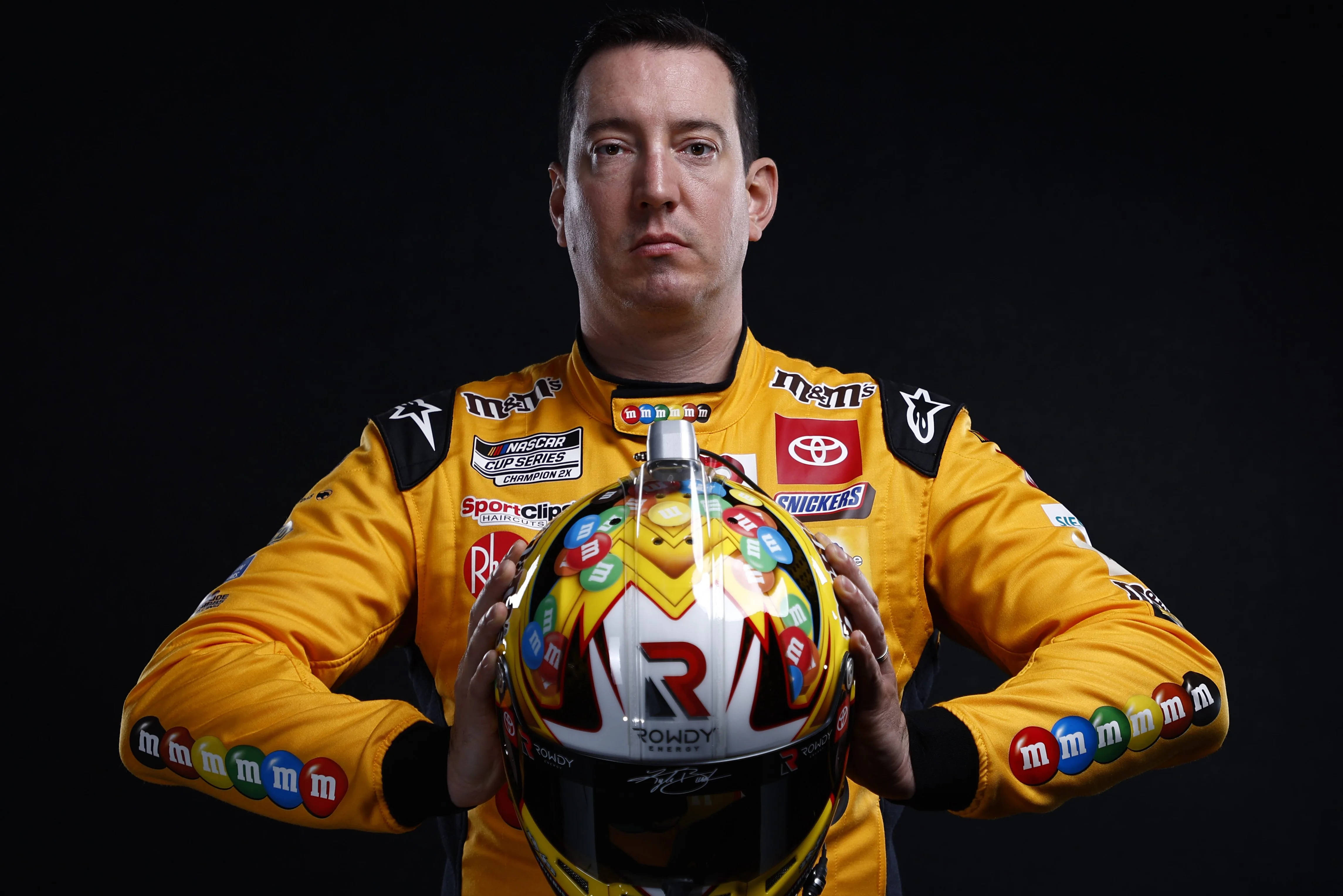 Photoshoot 桌布 壁紙 — 一位身穿亮黃色M&M's品牌賽車服的NASCAR車手，在黑色背景前手持色彩繽紛的頭盔。