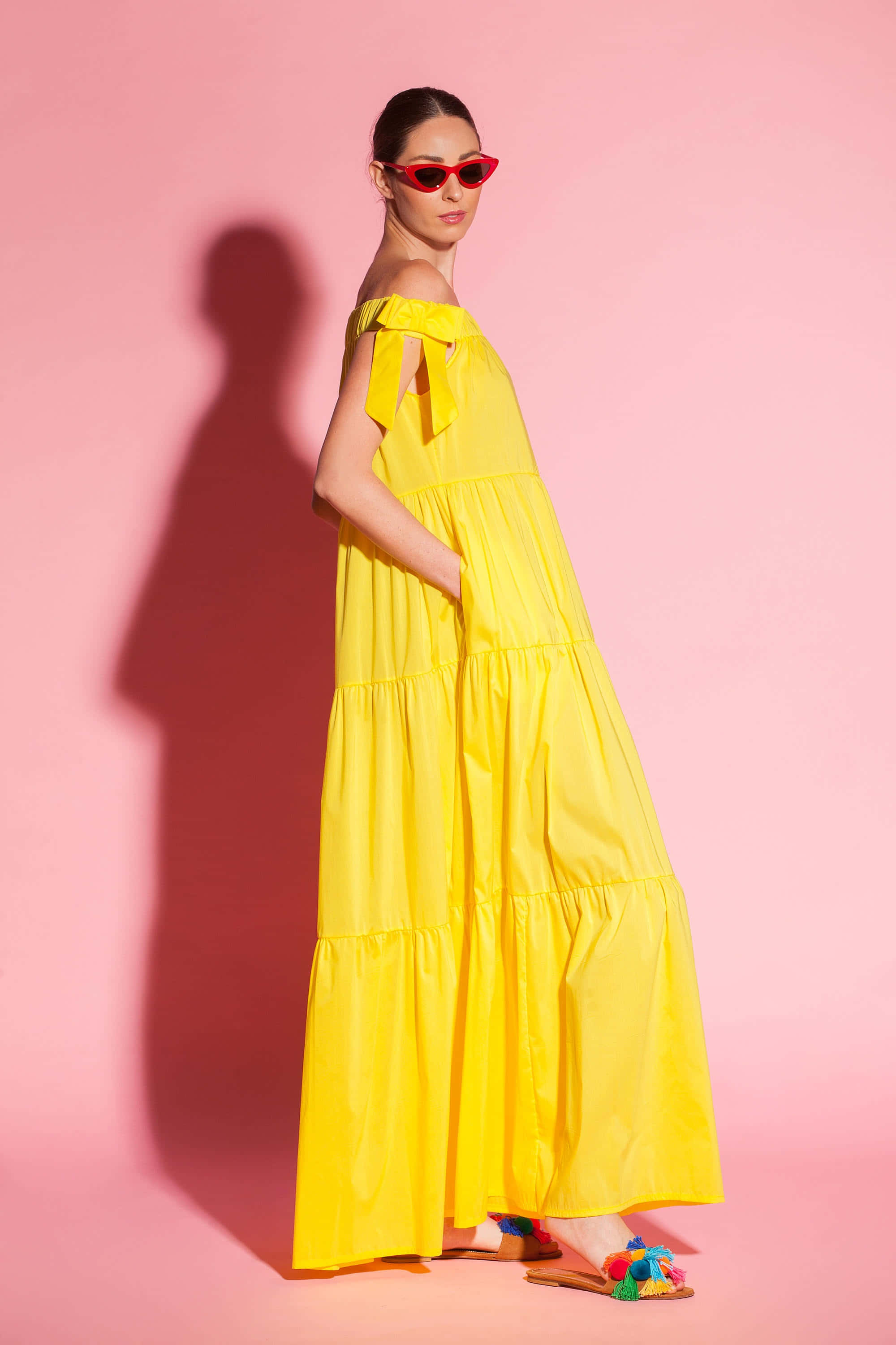 Yellow Fashion 桌布 壁紙 — 一位身穿亮黃色分層連衣裙和紅色太陽眼鏡的女性，在柔和的粉紅色工作室背景前擺姿勢，帶有強烈的陰影。