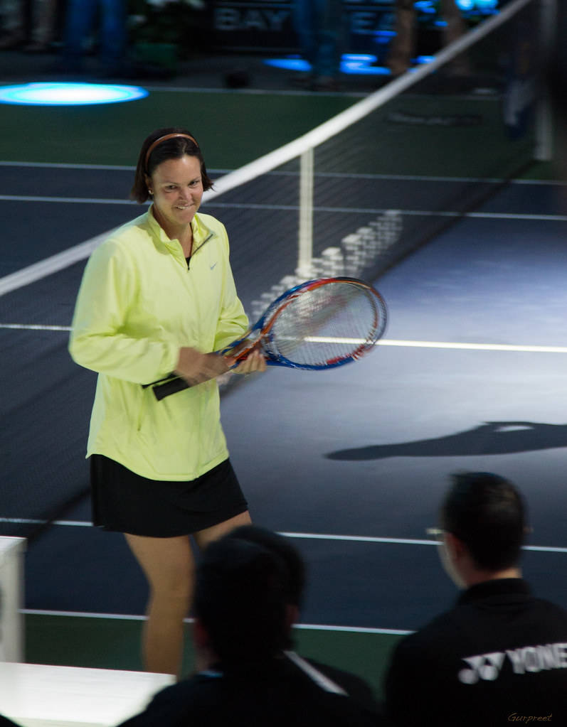 Tennis 桌布 壁紙 — 一位身穿亮黃色外套的女性網球選手在藍色燈光下的室內球場微笑著拿著球拍。