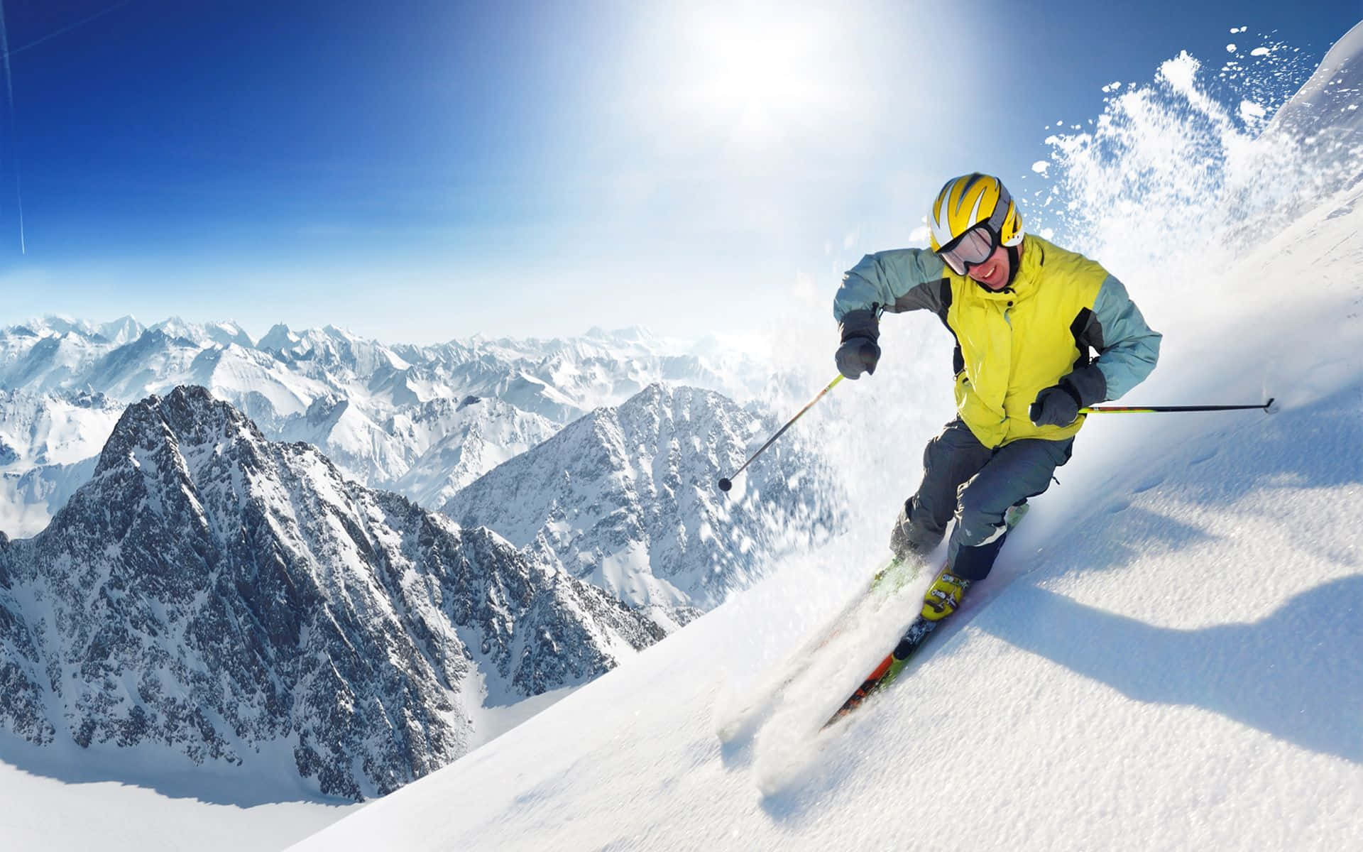 Winter Sports 桌布 壁紙 — 一位身穿亮黃色外套的滑雪者，在晴朗的藍天下，沿著白雪覆蓋的山坡滑下，身後濺起粉雪。