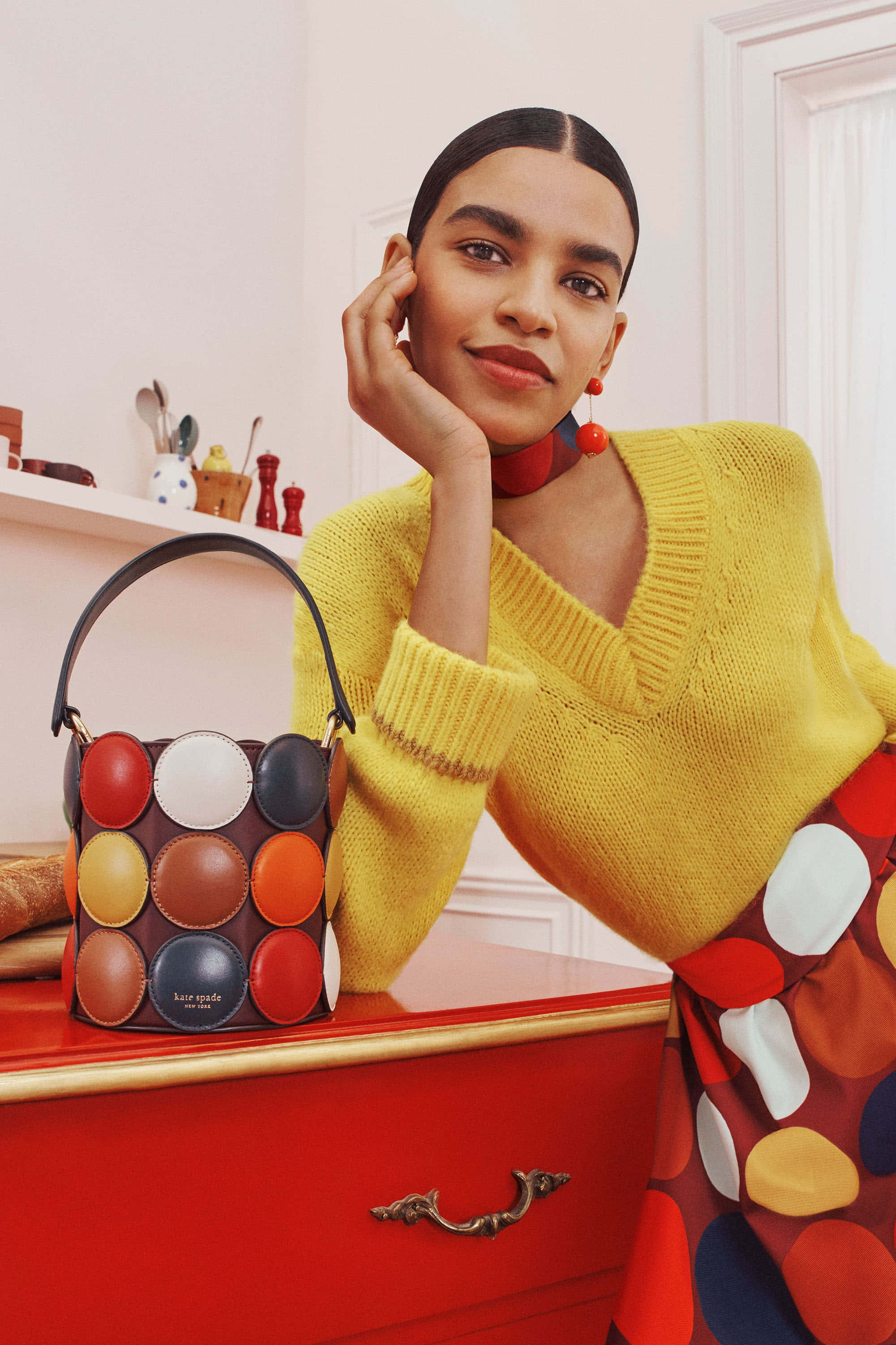 kate spade 桌布 壁紙 — 一位身穿亮黃色毛衣的女性，在光線柔和的房間裡，在紅色梳妝檯上一個色彩繽紛的圓點手提包旁擺姿勢。