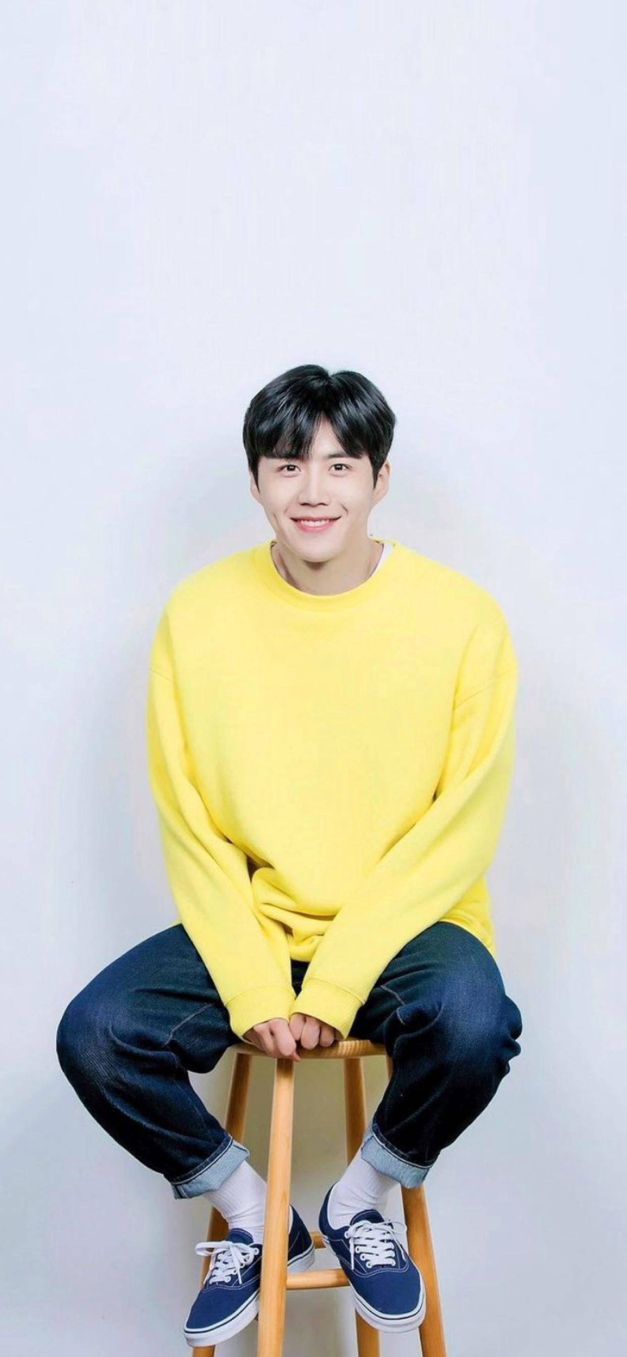 Kim Seon Ho 桌布 壁紙 — 一位身穿亮黃色毛衣的年輕男子坐在白色背景前的木凳上微笑。