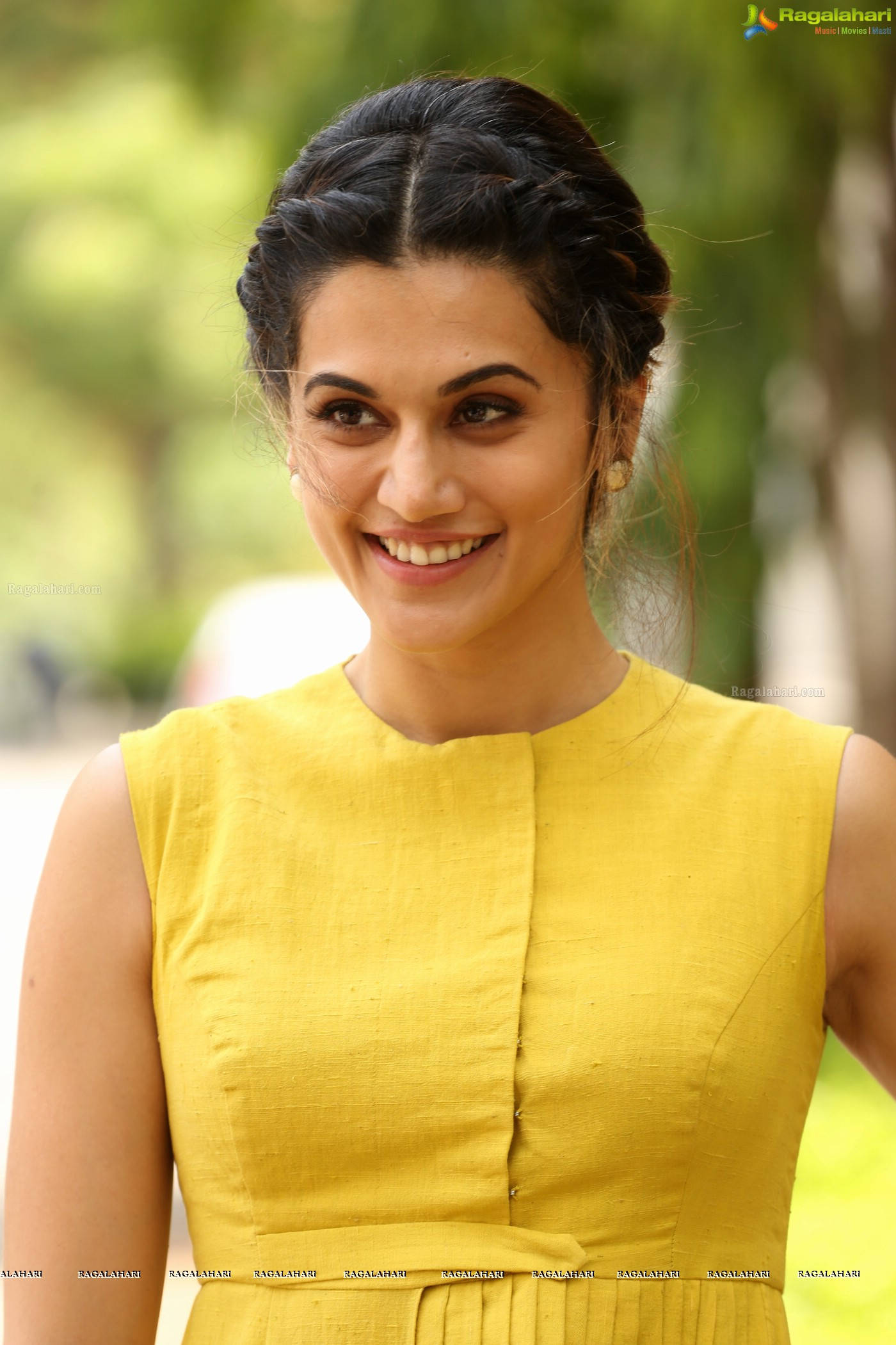 Taapsee Pannu 桌布 壁紙 — 一位身穿亮黃色無袖連衣裙的女性在柔和的綠色戶外背景下溫暖地微笑。