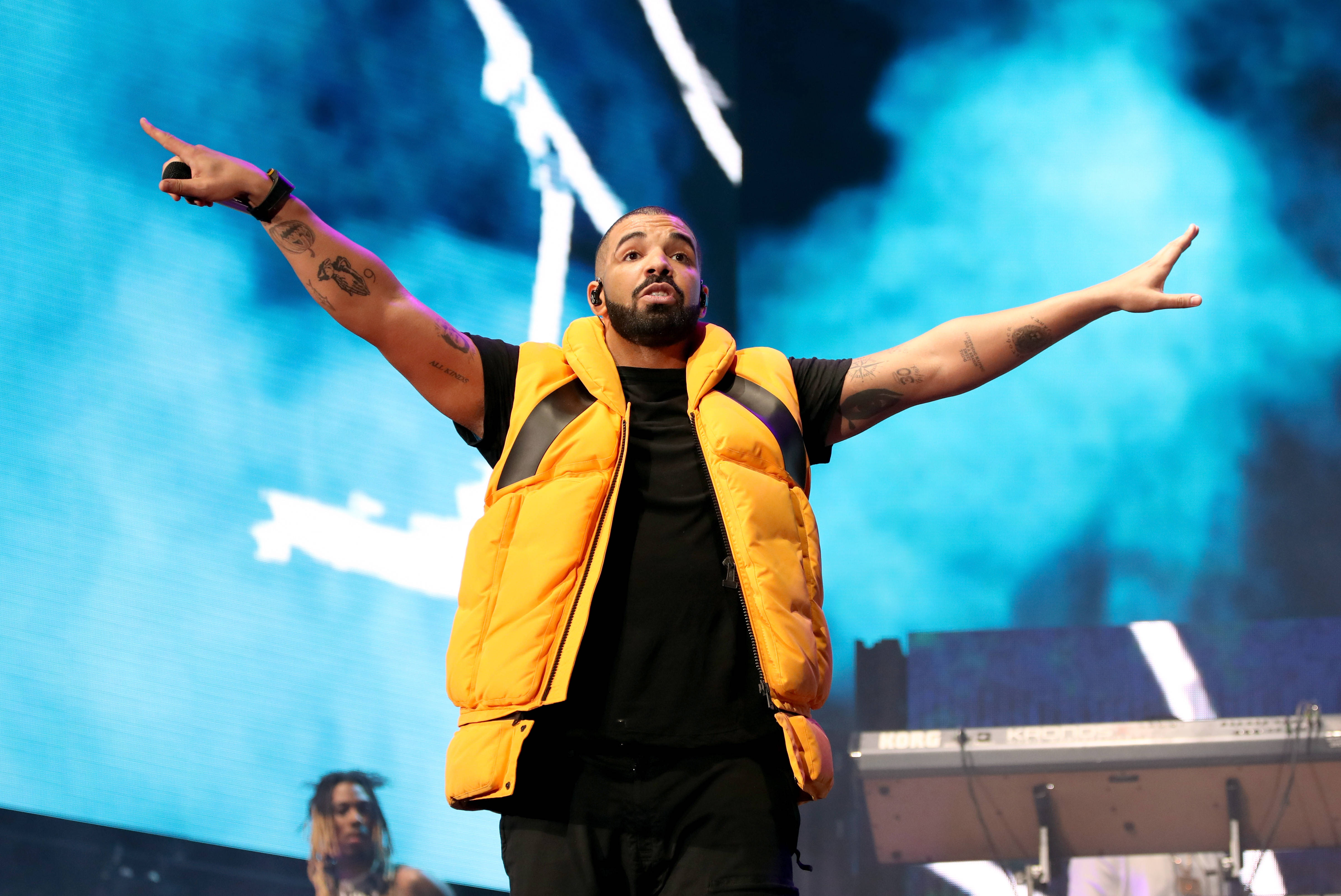 Drake 桌布 壁紙 — 一位身穿亮黃色背心的男性表演者在藍色燈光和煙霧效果的舞台上充滿活力地做手勢。