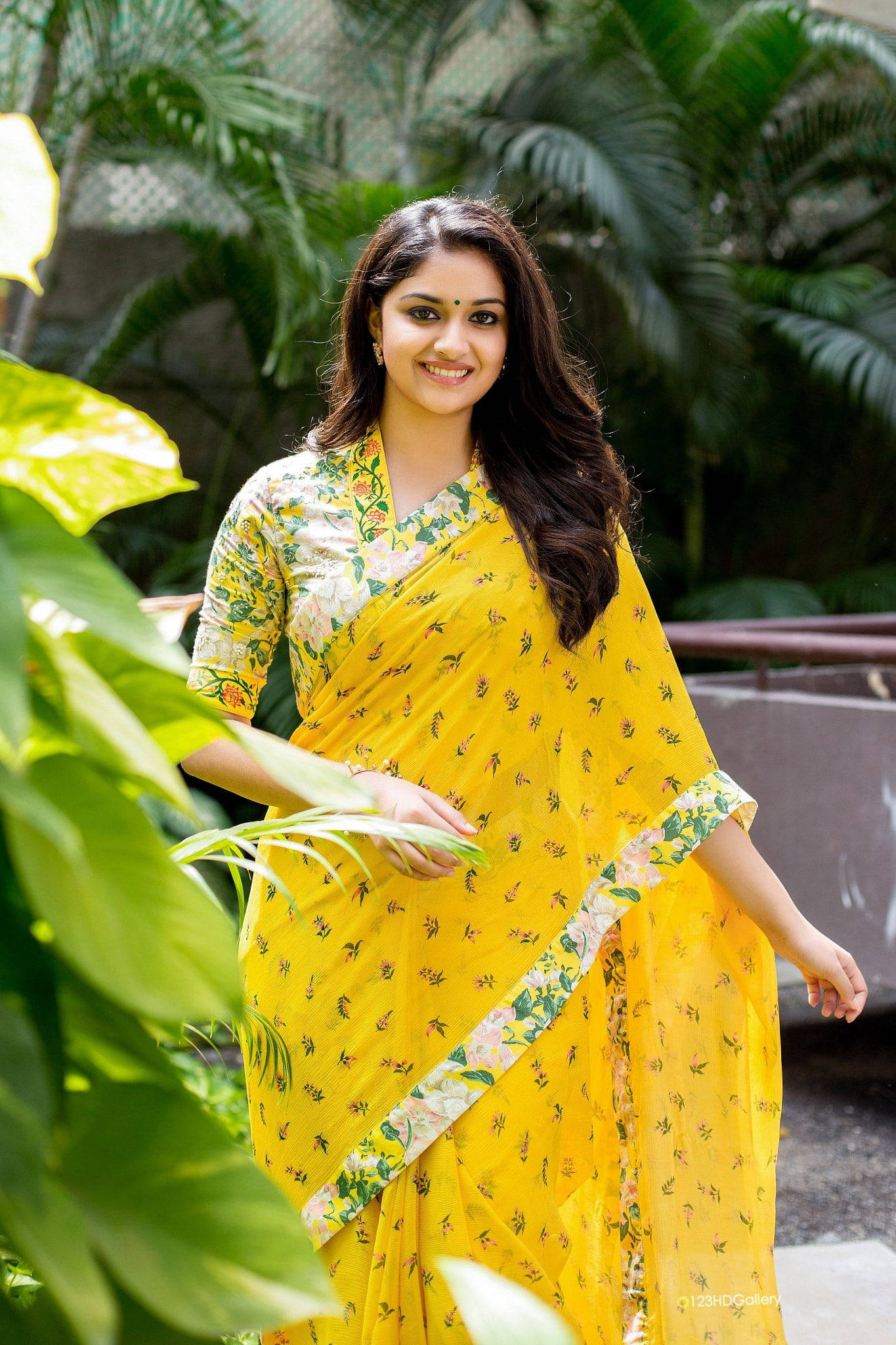 telugu 桌布 壁紙 — 一位身穿亮黃色花卉紗麗的女性在茂密的綠色熱帶植物中溫暖地微笑。