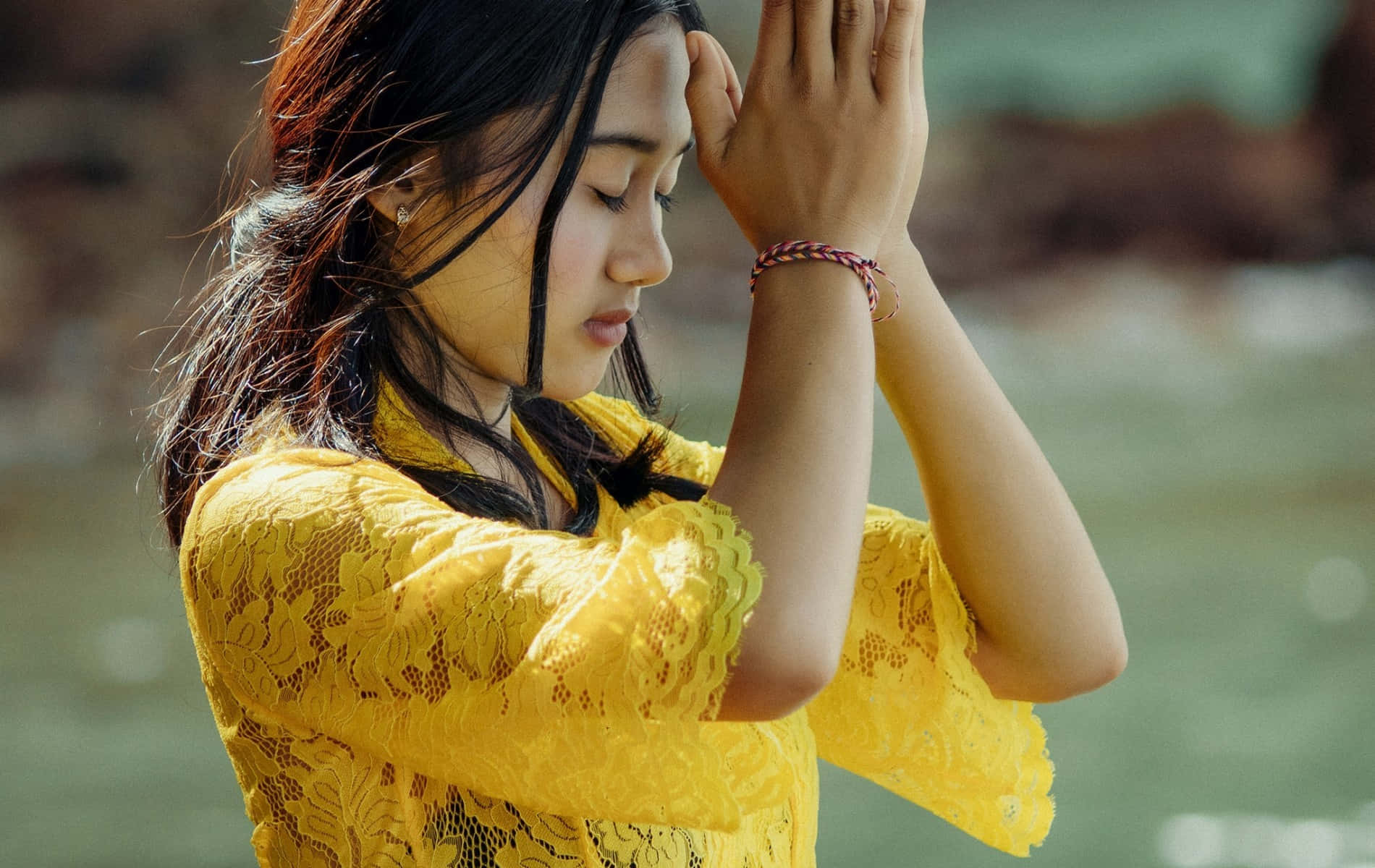 Praying 桌布 壁紙 — 一位身穿亮黃色蕾絲上衣的年輕女性站在水邊，雙手在臉龐附近交握，沉浸在靜謐的沉思時刻。
