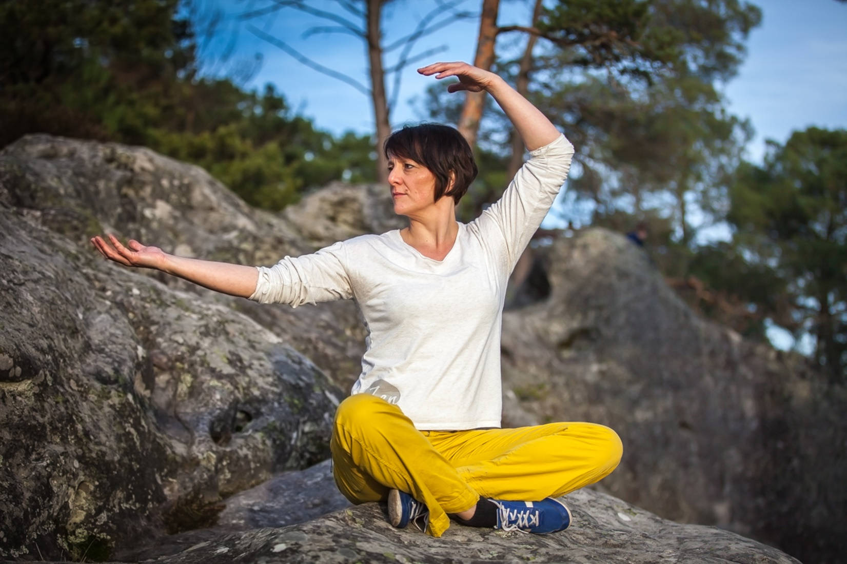 Exercise 桌布 壁紙 — 一位身穿亮黃色褲子的女性，在森林中的岩石露頭上盤腿而坐，雙臂優雅地伸展成瑜伽姿勢，背景是清澈的藍天。