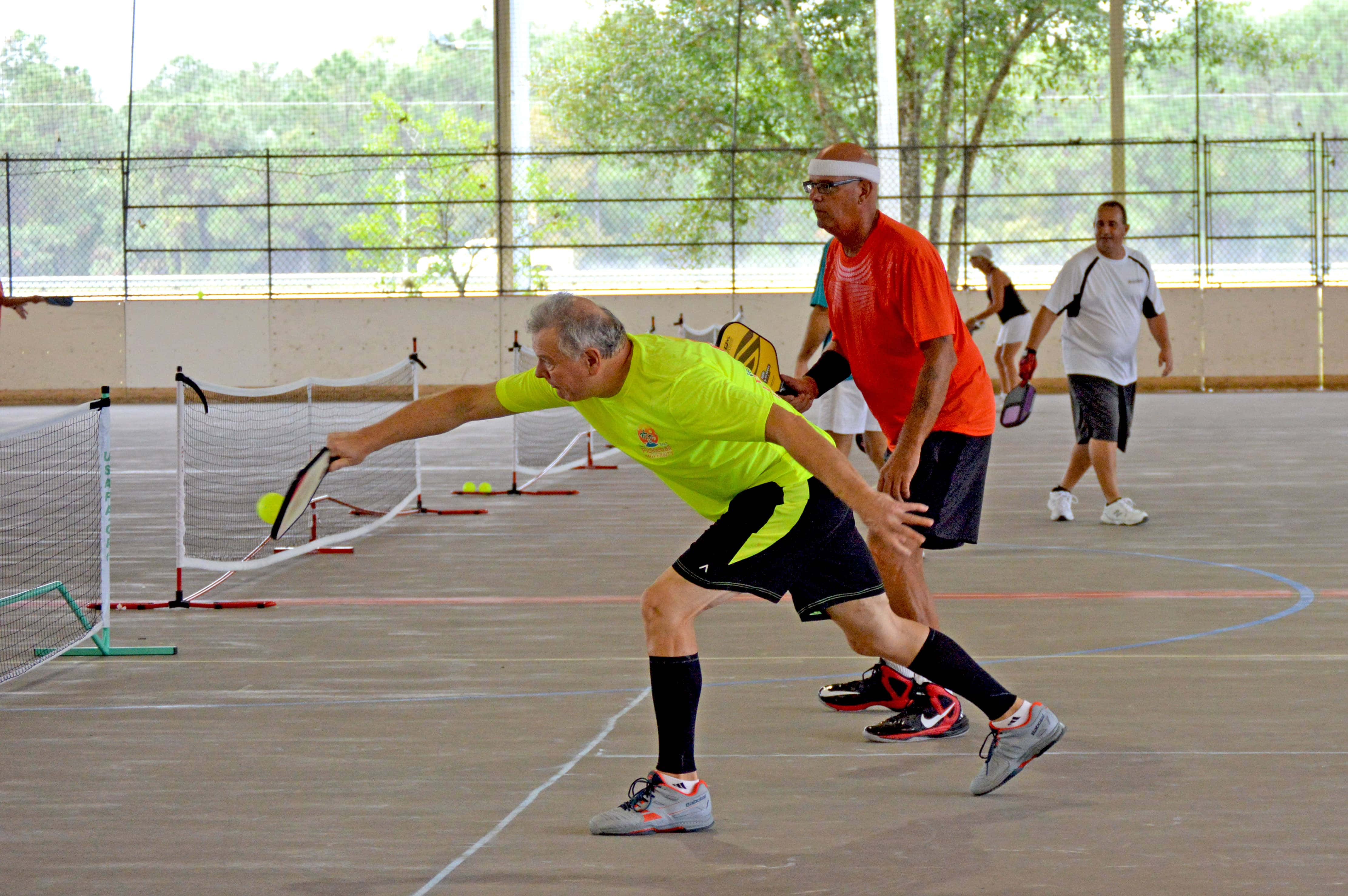 Pickleball 桌布 壁紙 — 一位身穿亮黃色襯衫的年長男性在室內比賽中撲向一個匹克球，與其他球員對戰。