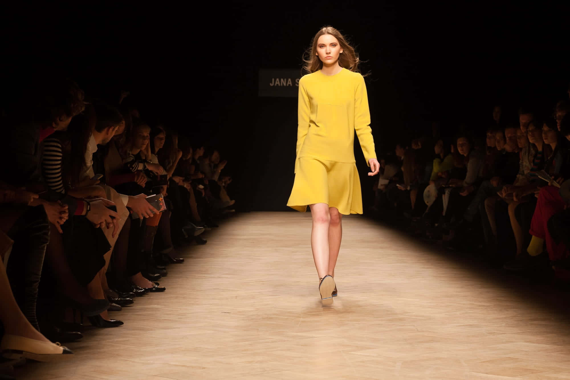 Runway 桌布 壁紙 — 一位身穿亮黃色連衣裙的模特兒自信地走在燈光昏暗的伸展台上，兩旁坐著觀眾。
