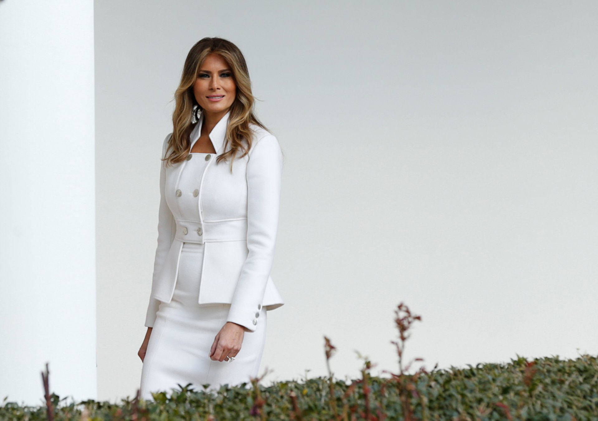 Melania 桌布 壁紙 — 一位身穿俐落白色訂製西裝的女性，自信地站在極簡白色牆壁前，腳邊有綠植。